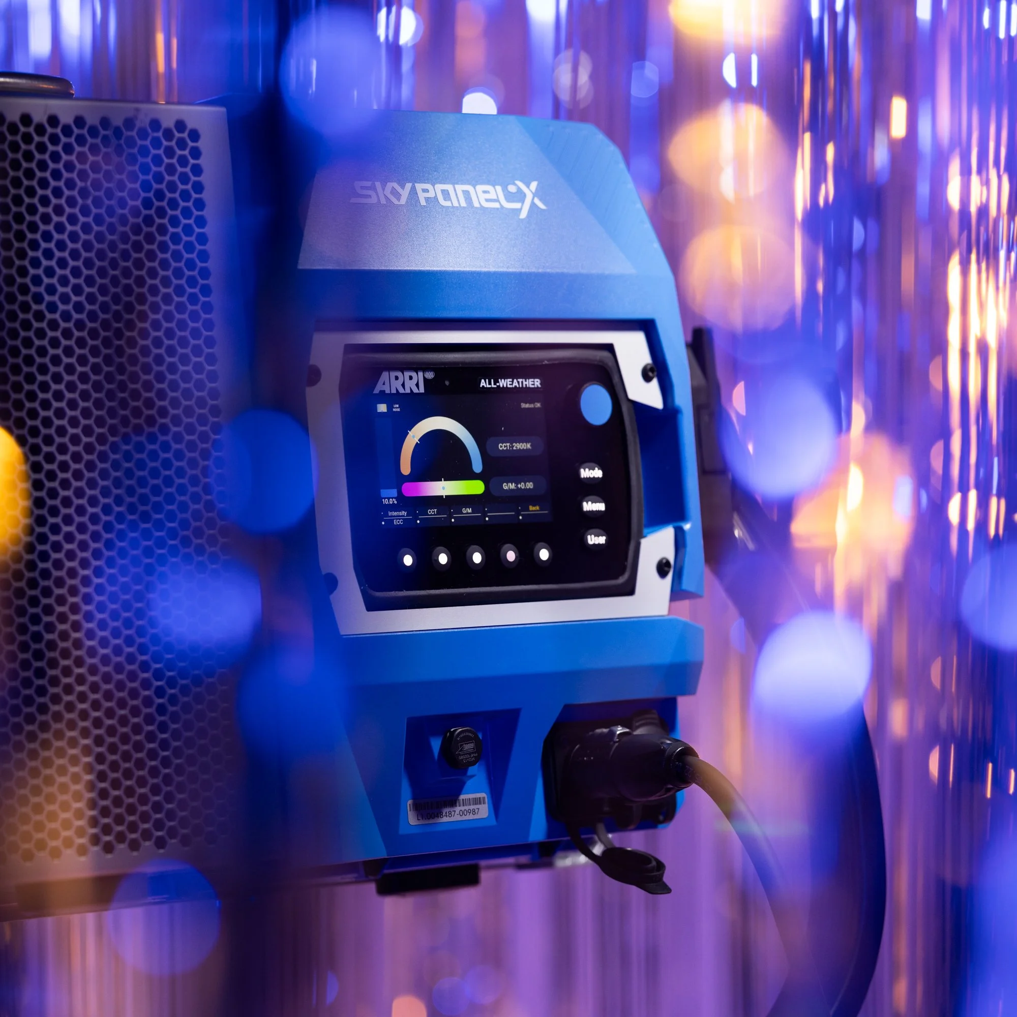 A close-up of a blue ARRI SkyPanelX LED light with a digital control screen displaying weather and color temperature settings, mounted on a stand with colorful bokeh lights in the background.