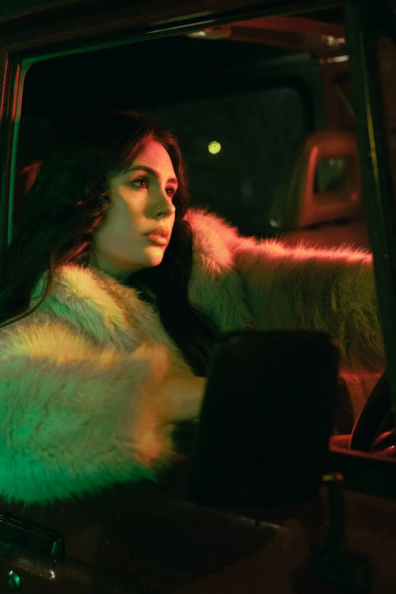 A woman with long dark hair and a nose piercing wearing a fur coat sitting in the driver's seat of a vehicle at night, illuminated by colored lights outside.