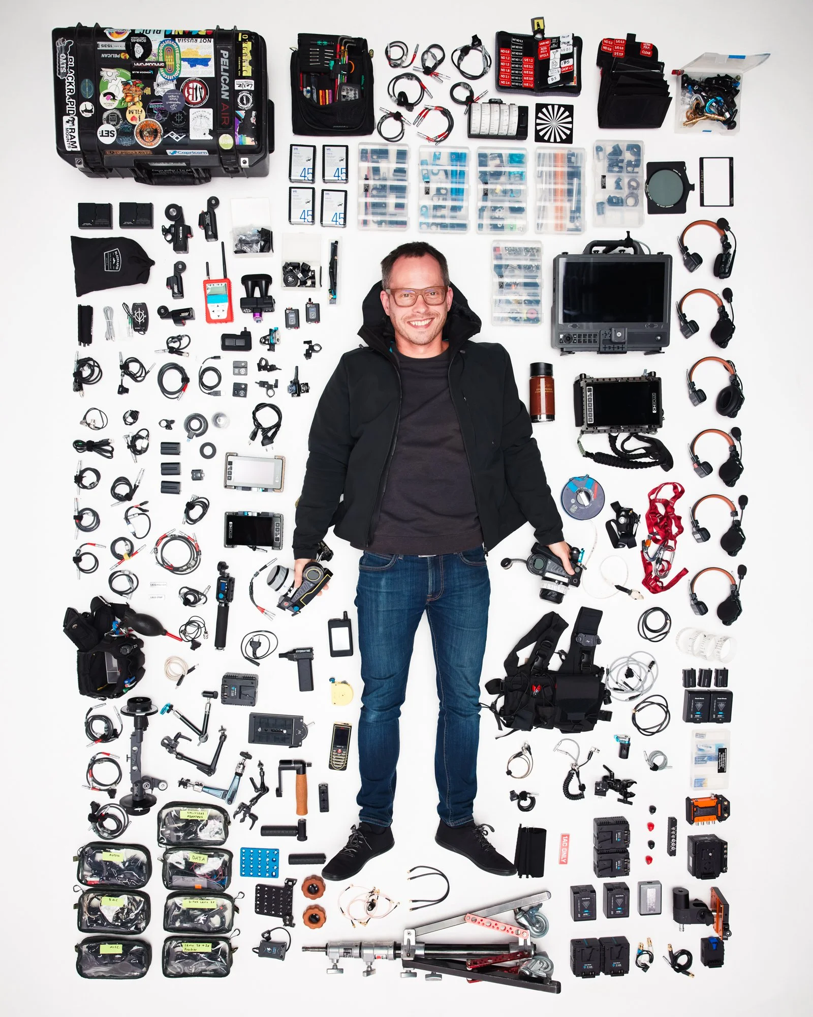 A man lying on the floor surrounded by various photography and videography equipment, including cameras, lenses, cables, batteries, and accessories.