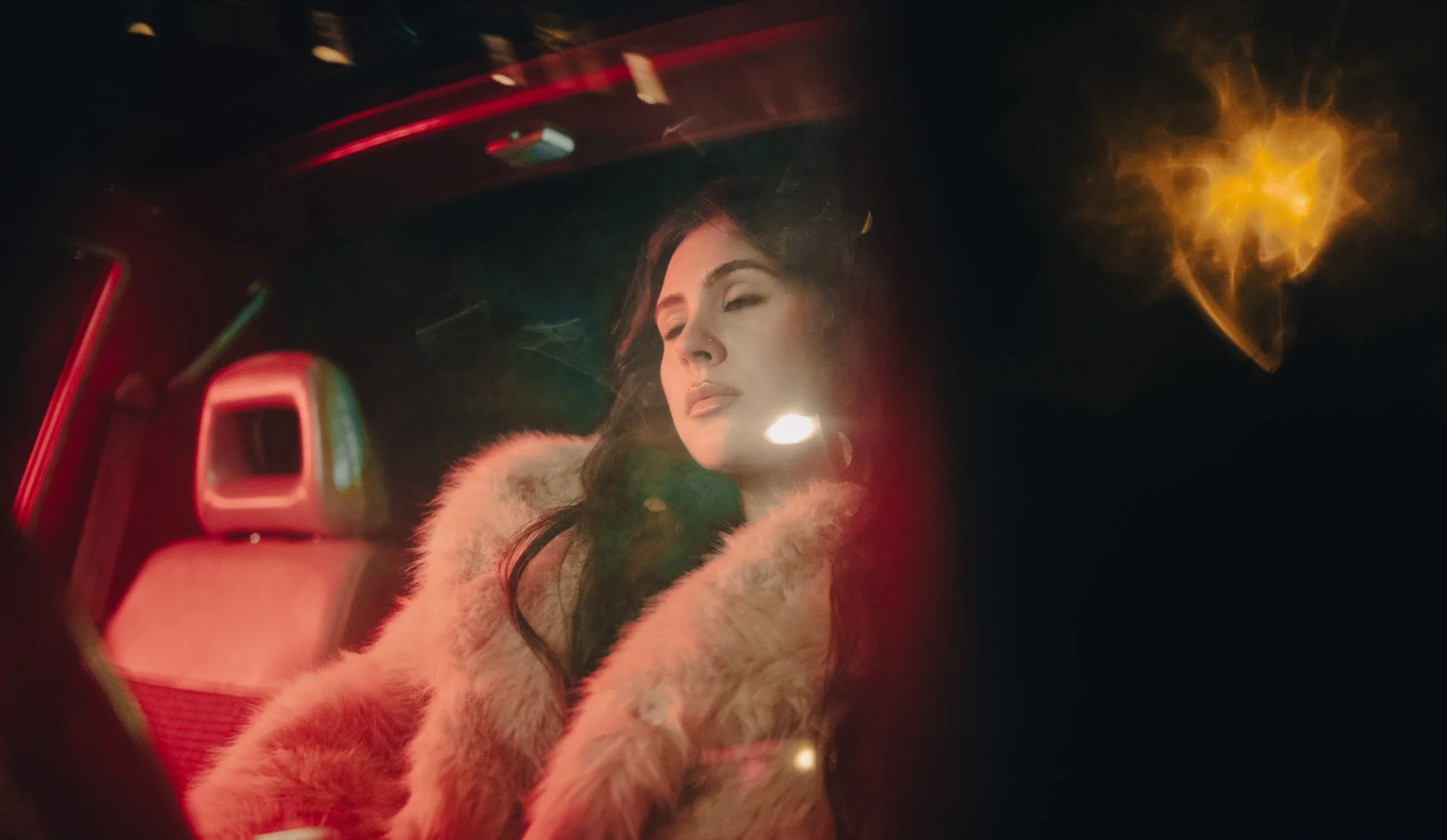 A woman with dark hair, wearing a fur coat, is sitting in a car at night with her eyes closed, illuminated by colorful, blurred neon lights outside.