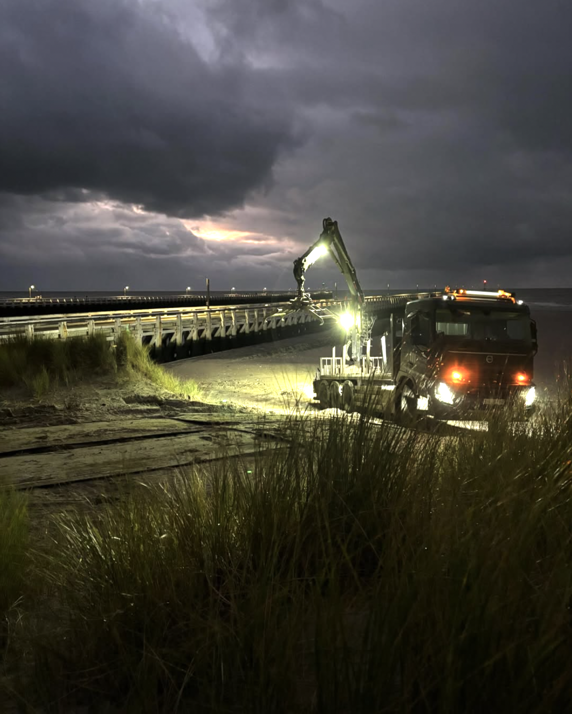 Een vrachtwagen met een graafmachine aan het werk op een strand bij schemering of 's nachts, met donkere wolken en een bliksemlicht op de horizon.