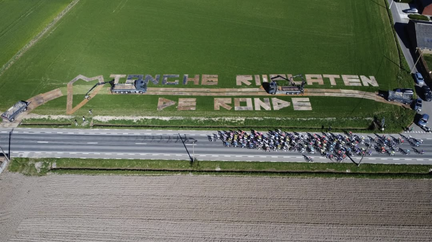 Luchtfoto van een groene grasweide met tekst gemaakt van planten of materialen, dat zegt 'TENGHE NUIATEEN BE RONDE'. Er zijn voertuigen die de tekst aan het maken zijn, en een groep mensen en fietsen onder een brug aan de weg.