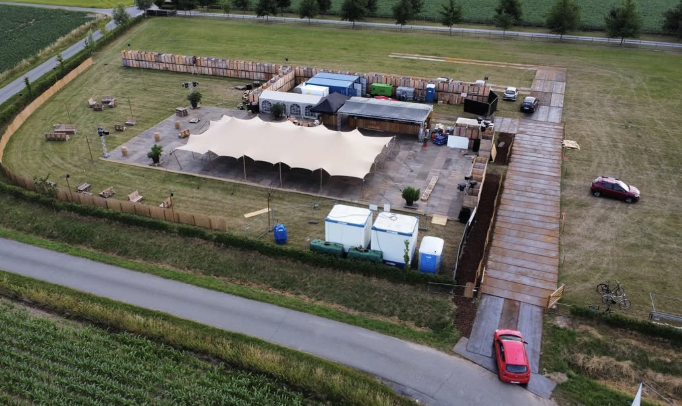 Een open veld met een grote witte tent en bouwmaterialen, omringd door een houten hekwerk en een pad met enkele auto's en fietsen.