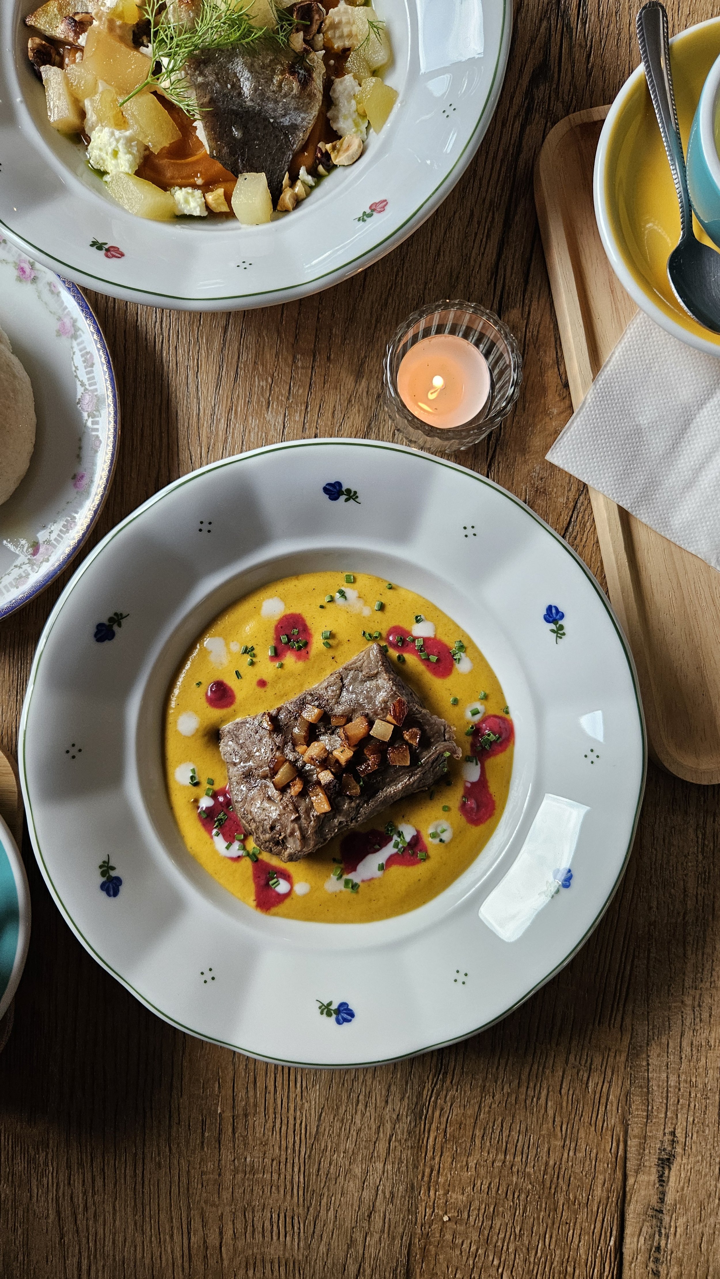 Gourmet dish on a decorated plate with meat and colorful sauce. Background includes another dish, small candle, and napkin.