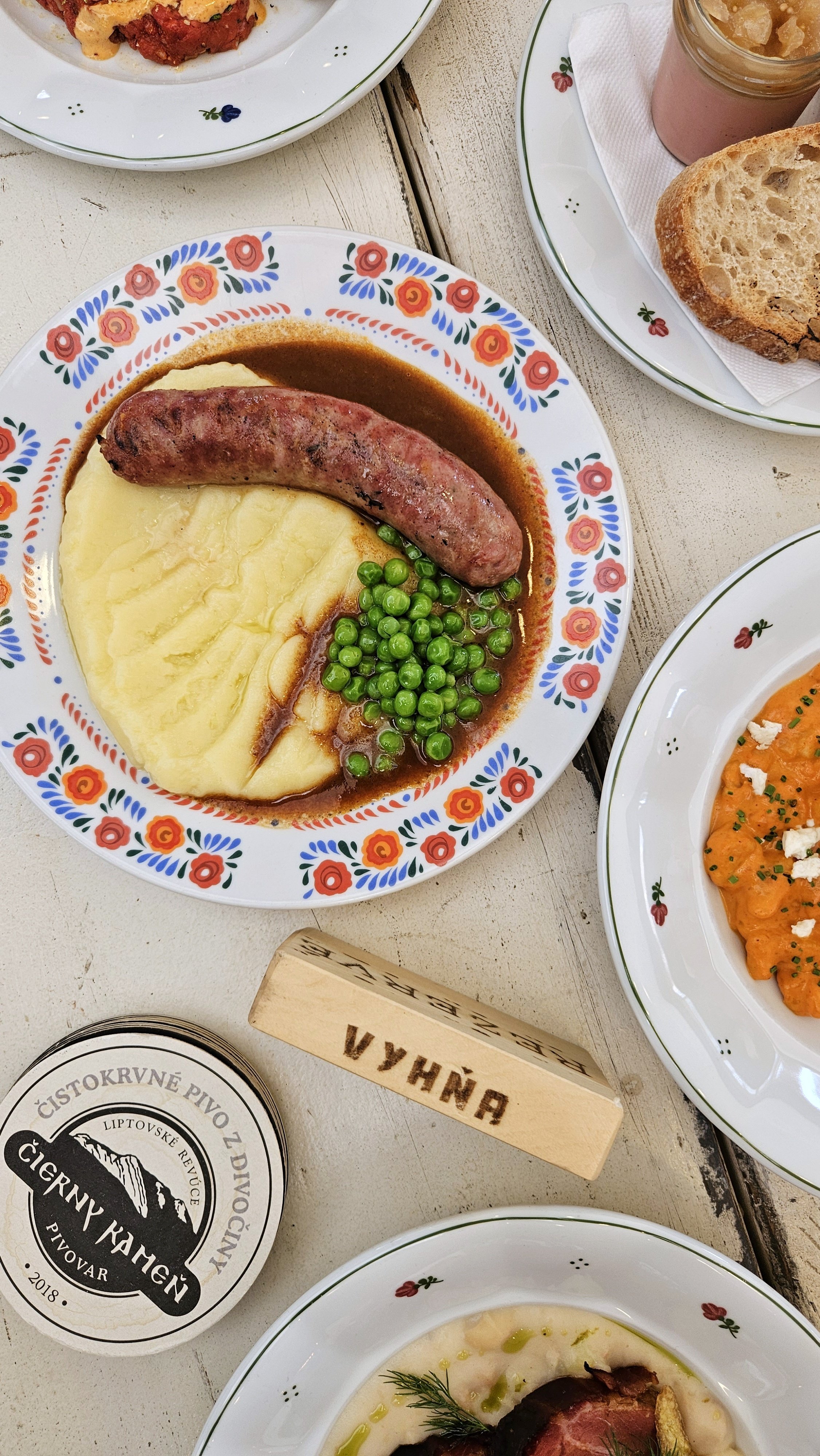 Plate with sausage, mashed potatoes, and peas, surrounded by other dishes and a beer coaster.