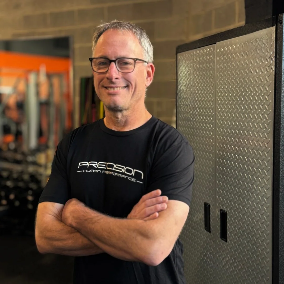 A smiling man with glasses and a beard in a gym, wearing a black t-shirt with 'Precision Human Performance' written on it, standing next to gym equipment.