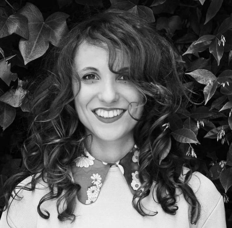 Portrait noir et blanc de l'artiste. Une femme souriante avec des cheveux bouclés devant un arrière-plan de feuilles. Son visage est lumineux.