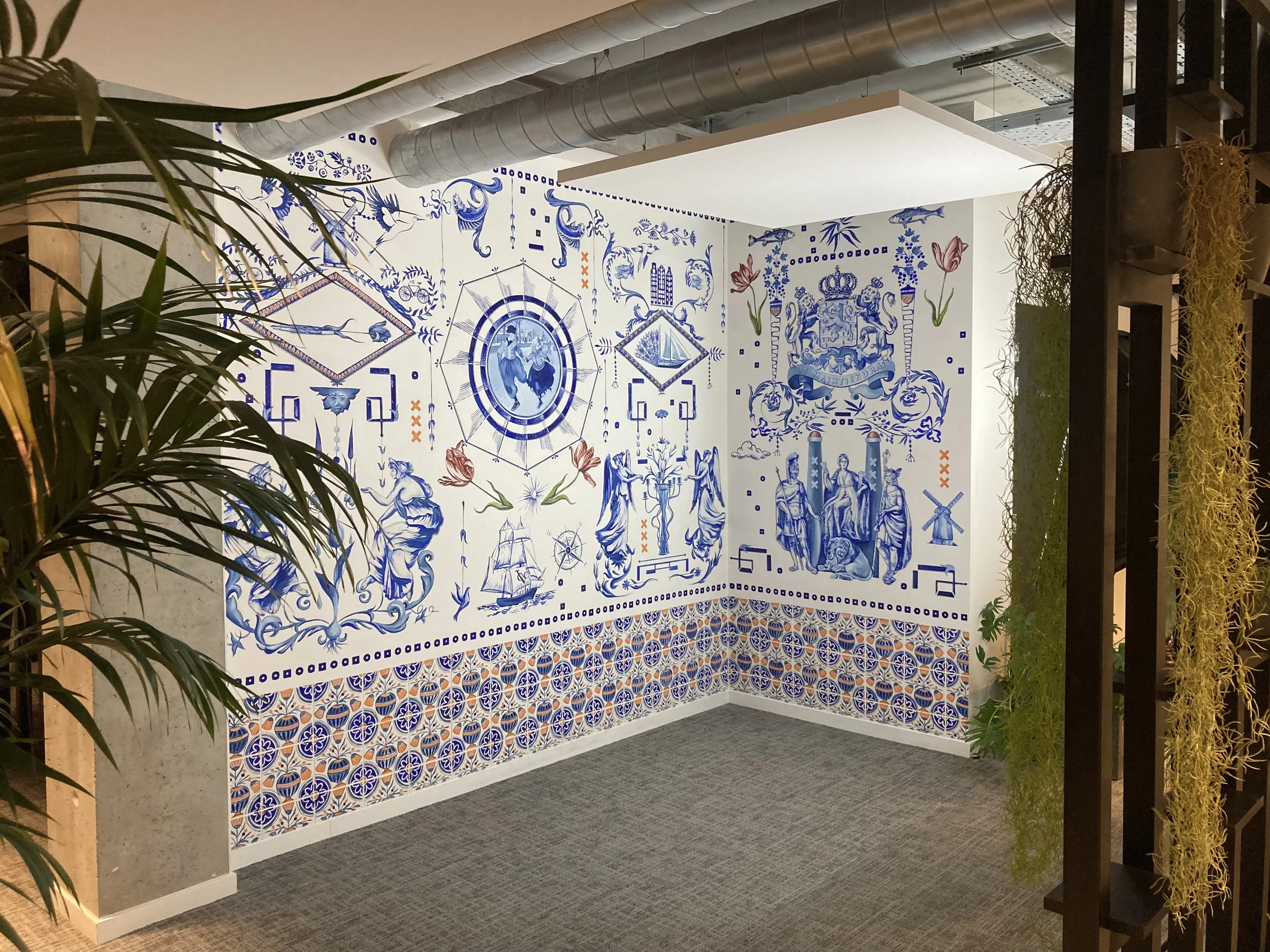 Une grande peinture recouvre un mur dans un espace moderne. On y voit des dessins en camaïeu de bleus qui évoquent la porcelaine de Delft, des objets et des symboles liés aux Pays-Bas, comme une histoire racontée sur les murs. Enduit minéral naturel.
