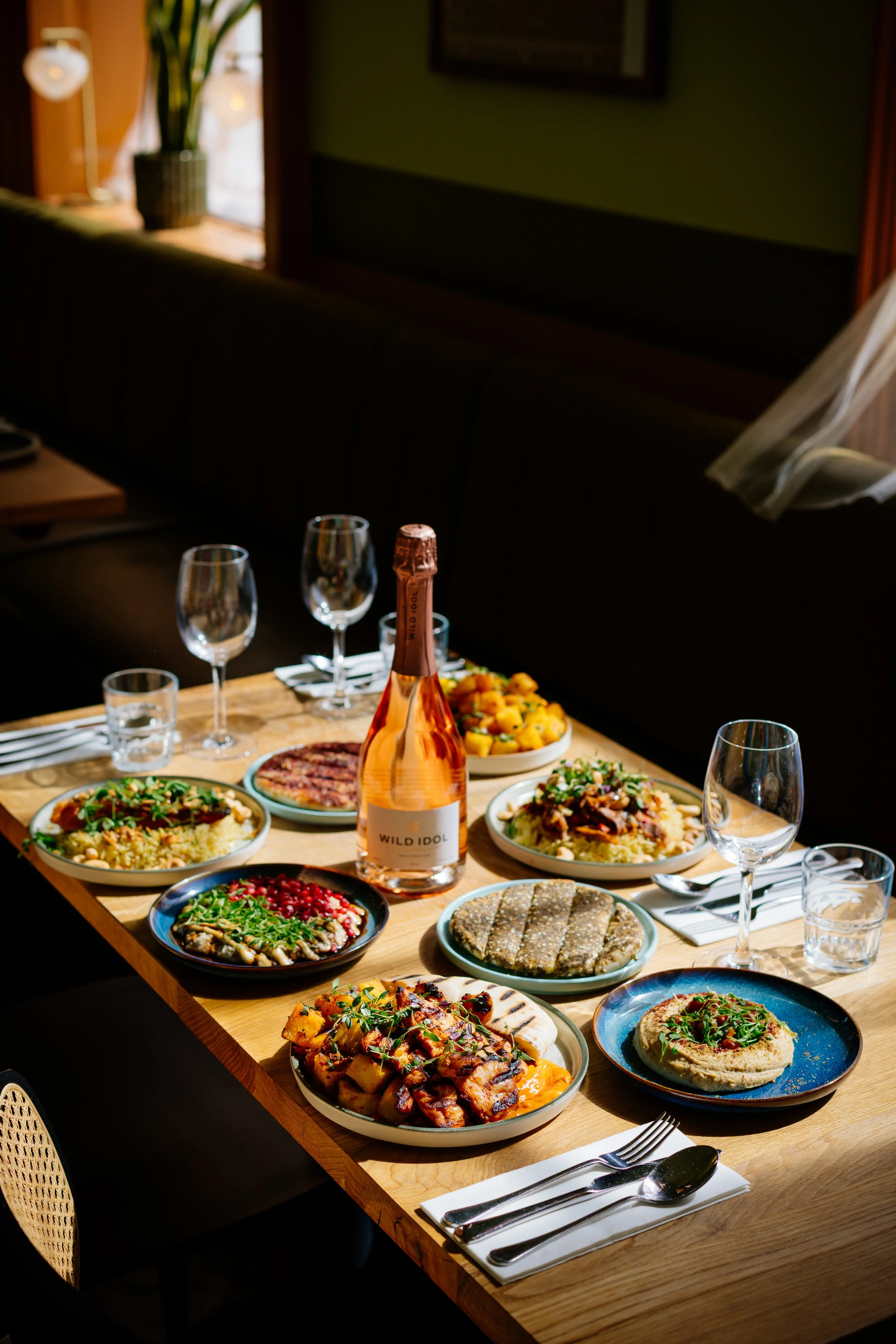 A wooden dining table set with various dishes including salads, grilled meats, and hummus, alongside wine glasses, a bottle of rosé wine, and cutlery, in a cozy restaurant with natural light and a bench seating area.