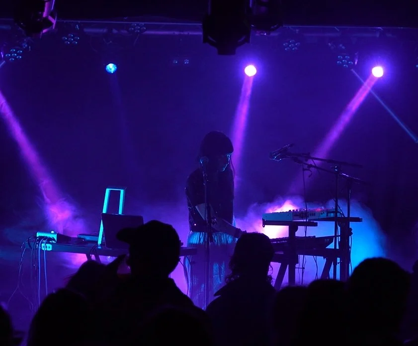 Une personne joue de la musique sur scène avec des lumières violettes et bleues, entourée de spectateurs.