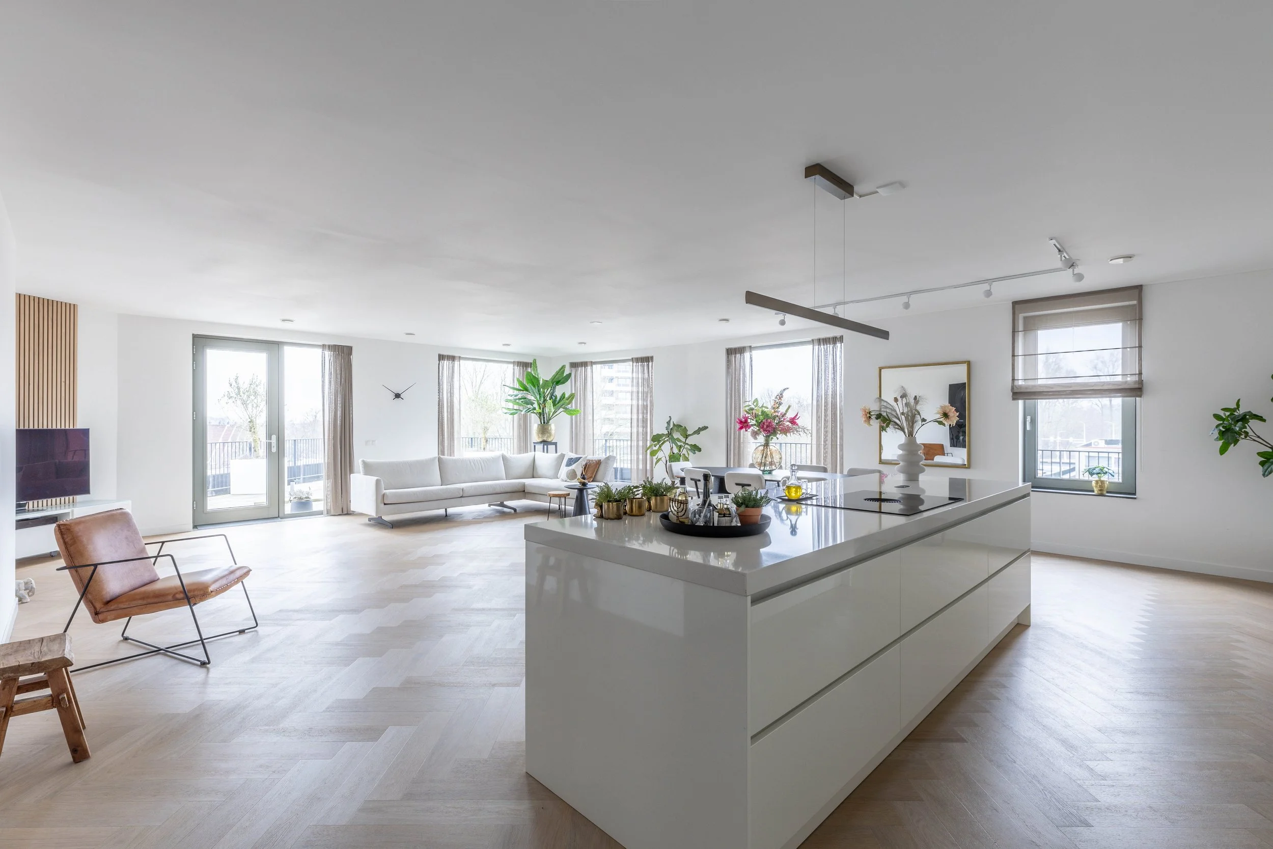 Lichte en ruime woonkamer met wit banken, grijze gordijnen, planten, en een witte keuken met bloemen, glaswerk en decoraties, grote ramen en houten vloer.
