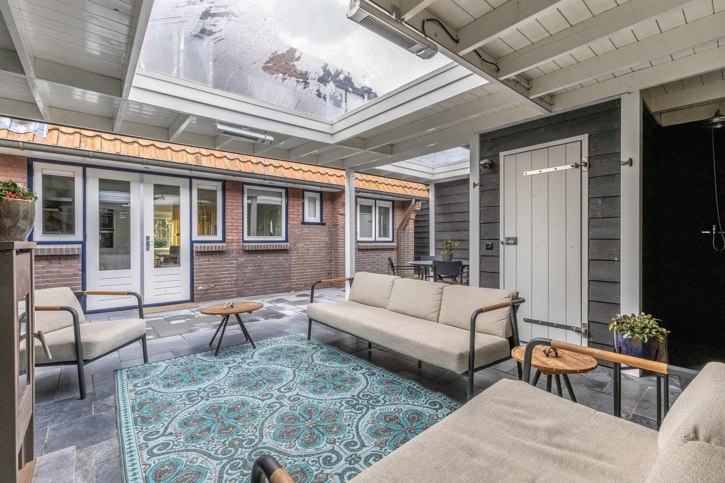 Overdekte buitenruimte met geregelde zitplaatsen, een blauwe vloerkleed, planten, een tafel en ramen met witte kozijnen aan een bakstenen muur.