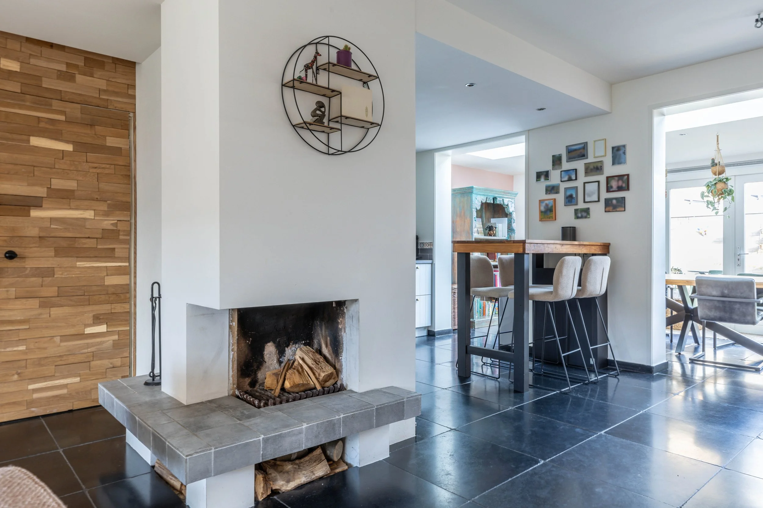 Interieur van een huis met een open haard, houten vloer, eetkamer en een muur met fotolijstjes.
