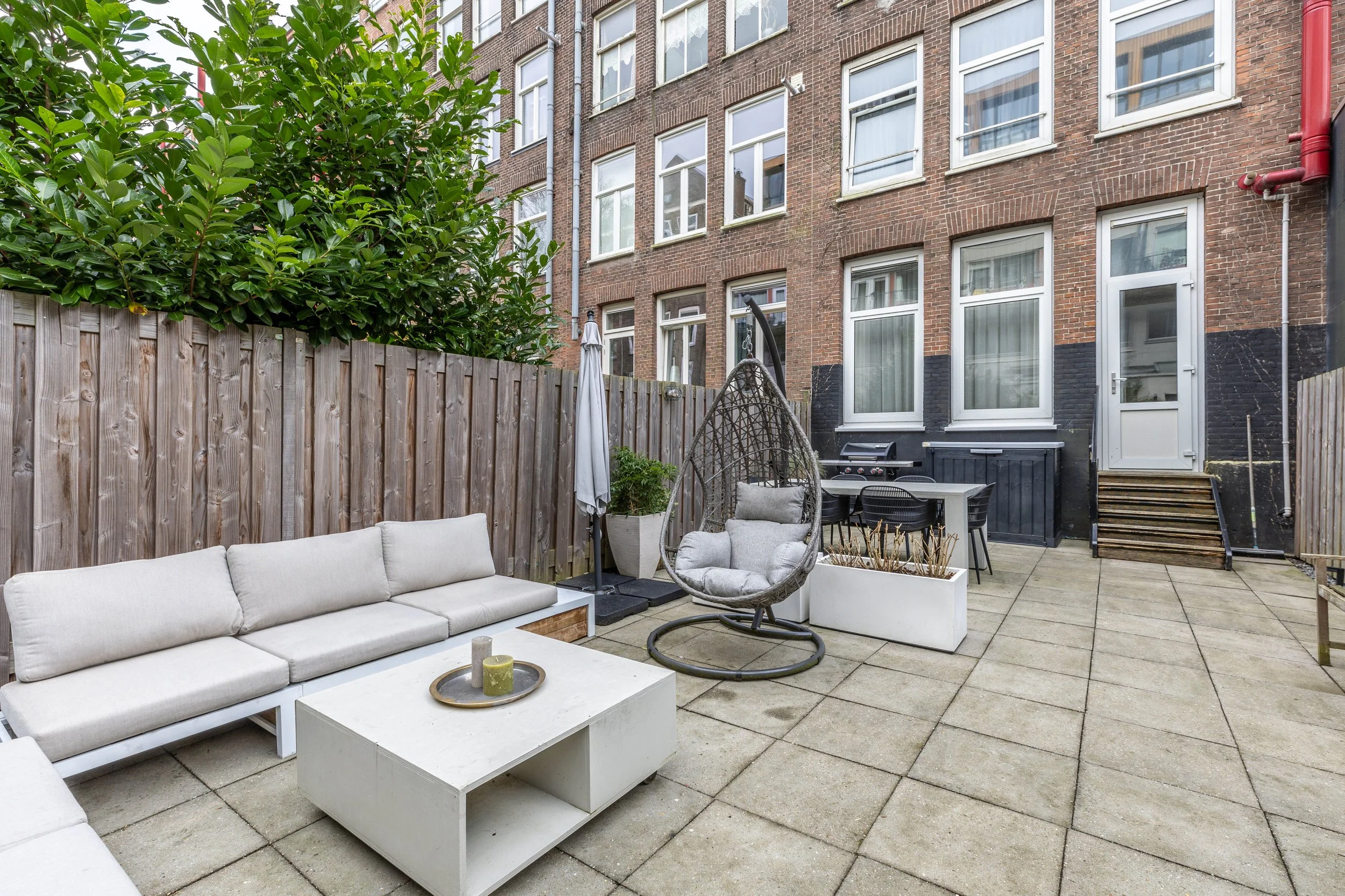 Een moderne buitenruimte met zithoek inclusief een witte bank, een witte salontafel met kaarsen, een hangstoel met kussens, een plafondparaplu, een barbecue en een eettafel met zes stoelen, omgeven door een houten schutting en uitzicht op een baksten