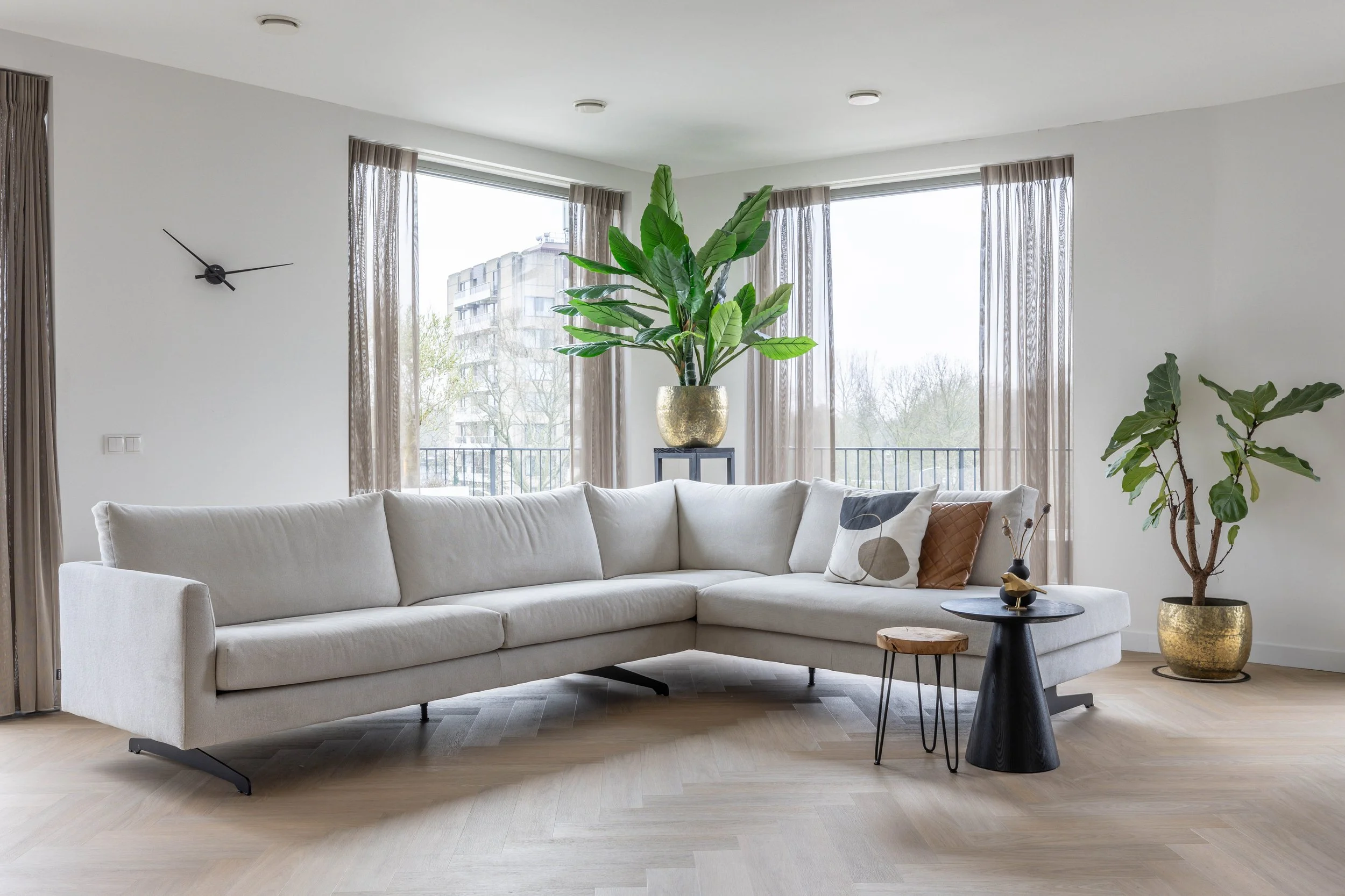 Woonkamer met een grote witte hoekbank, twee sierkussens (één met abstract patroon en één bruin), een houten bijzettafel, een zwart salontafeltje met decoratief object, grote glazen raampartijen met beige gordijnen, twee grote groene planten in goude