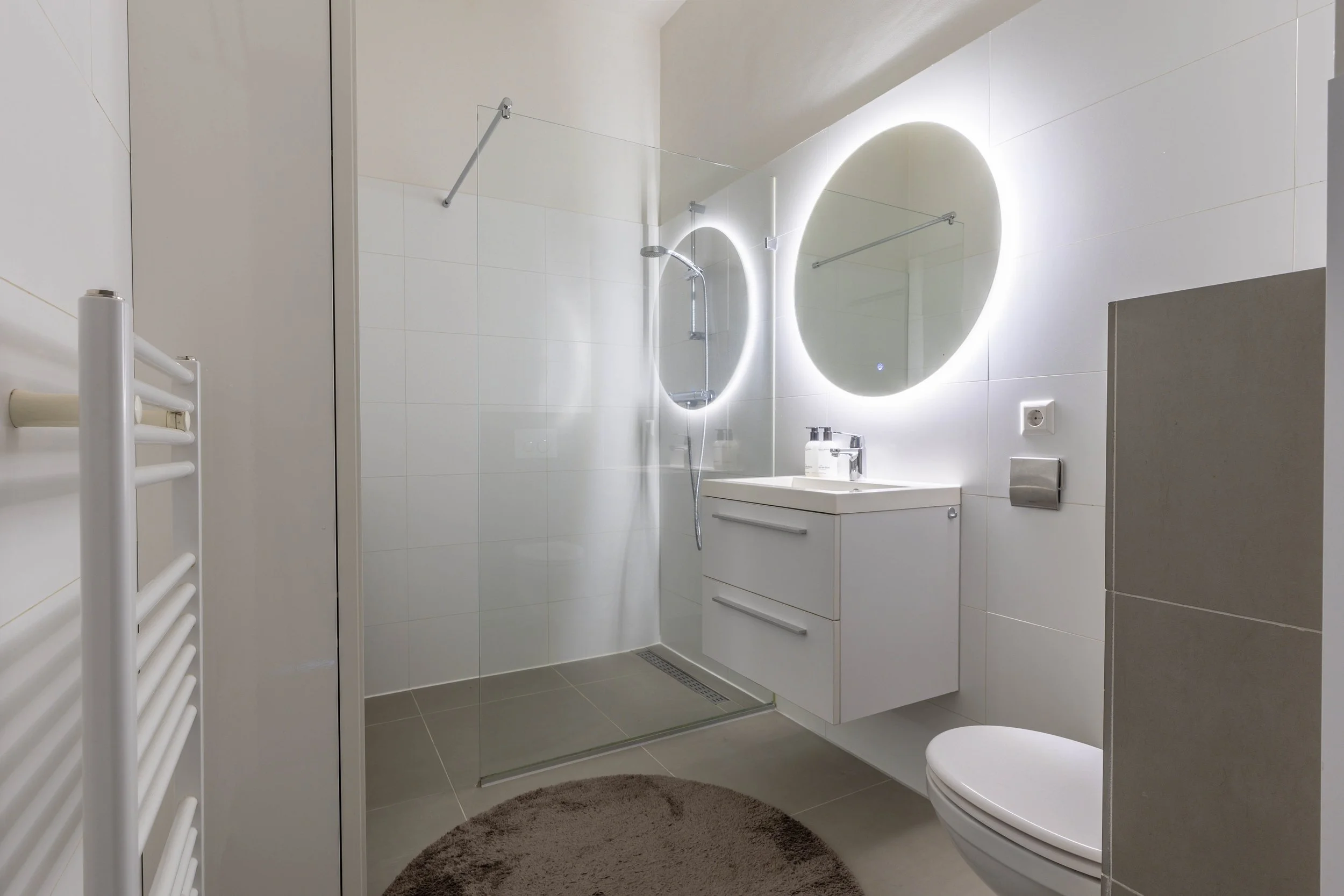 Een moderne, Minimalistische badkamer met wit tegelwand, inloopdouche met glaswand, ronde spiegel met LED-verlichting, witte badkamermeubel met wastafel, handdoekradiator, toilet en ronde badmat.