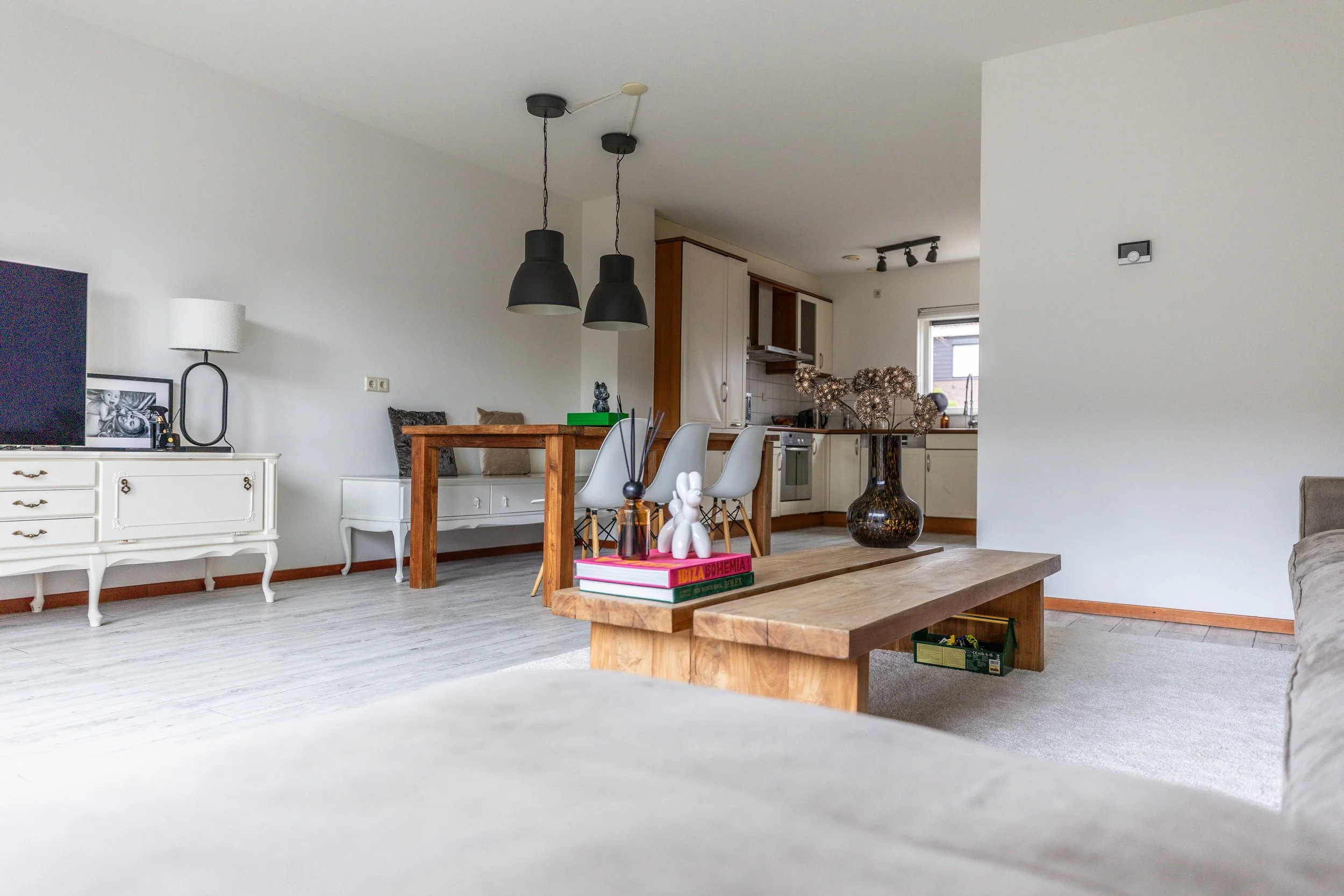 Woonkamer met witte muur, houten vloer, tv, witte kast, bank, eettafel met witte stoelen, keuken in achtergrond, bloemen, decoraties.