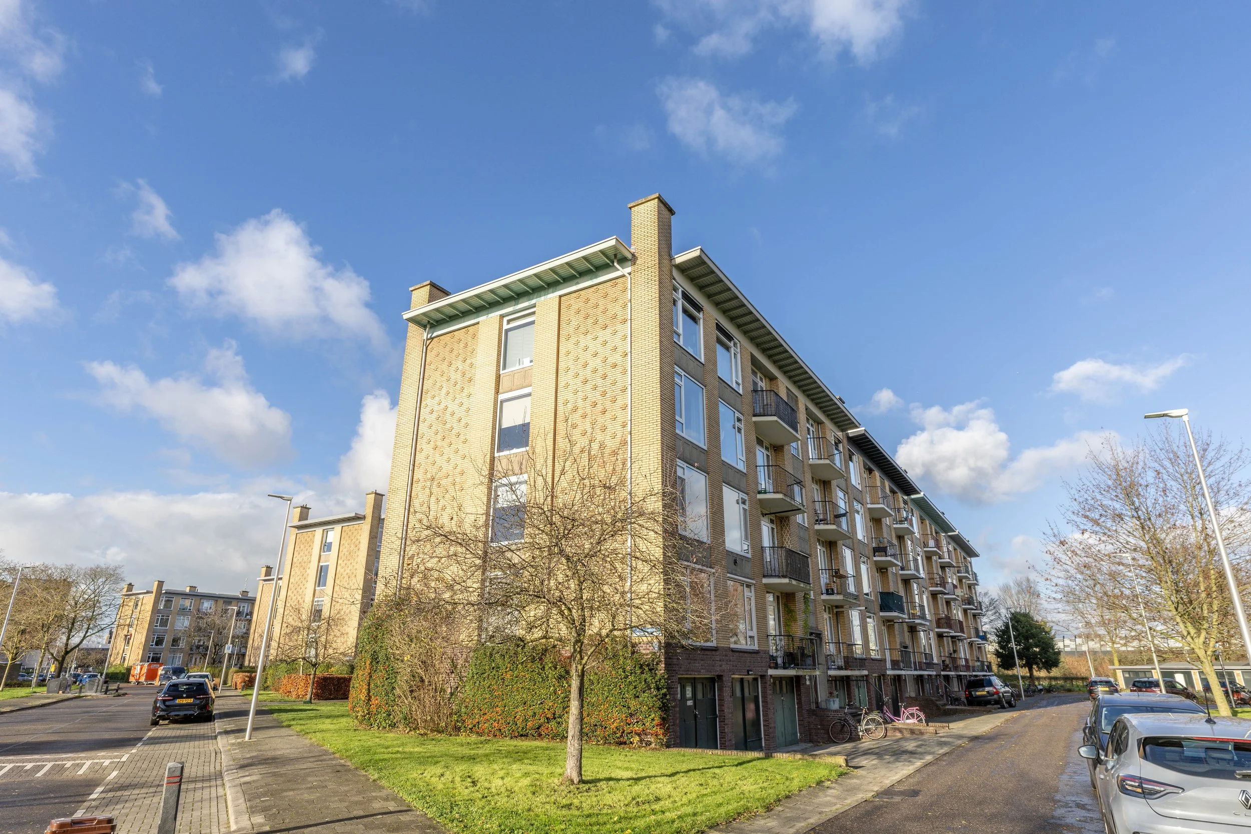Hoge appartementencomplex met meerdere verdiepingen, balkons, en een parkeerplaats met auto's, onder een blauwe sky met enkele wolken.