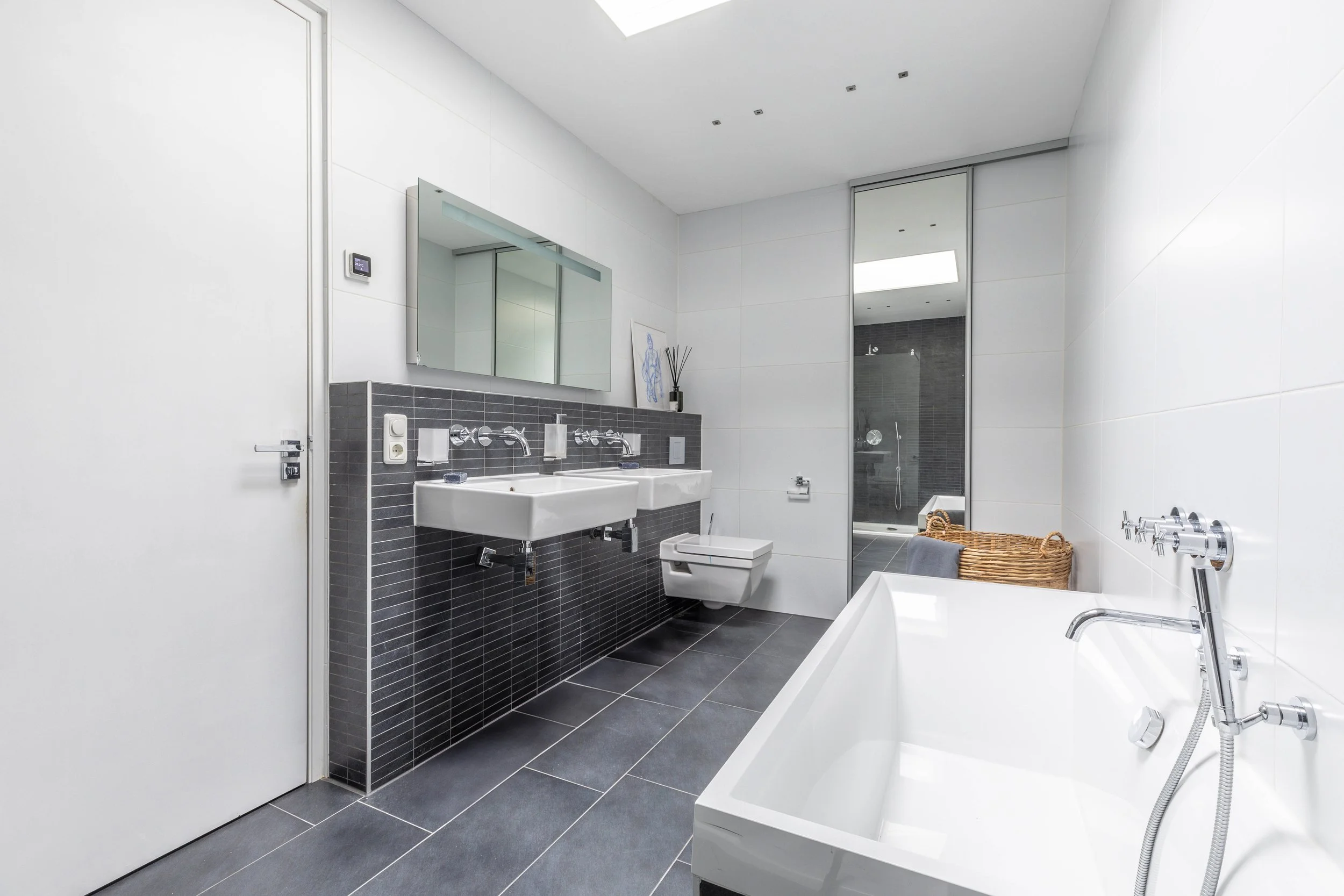 Moderne badkamer met dubbele wastafel, spiegel, wandcloset, een badkuip, een inloopdouche, en een wasmand met handdoeken.