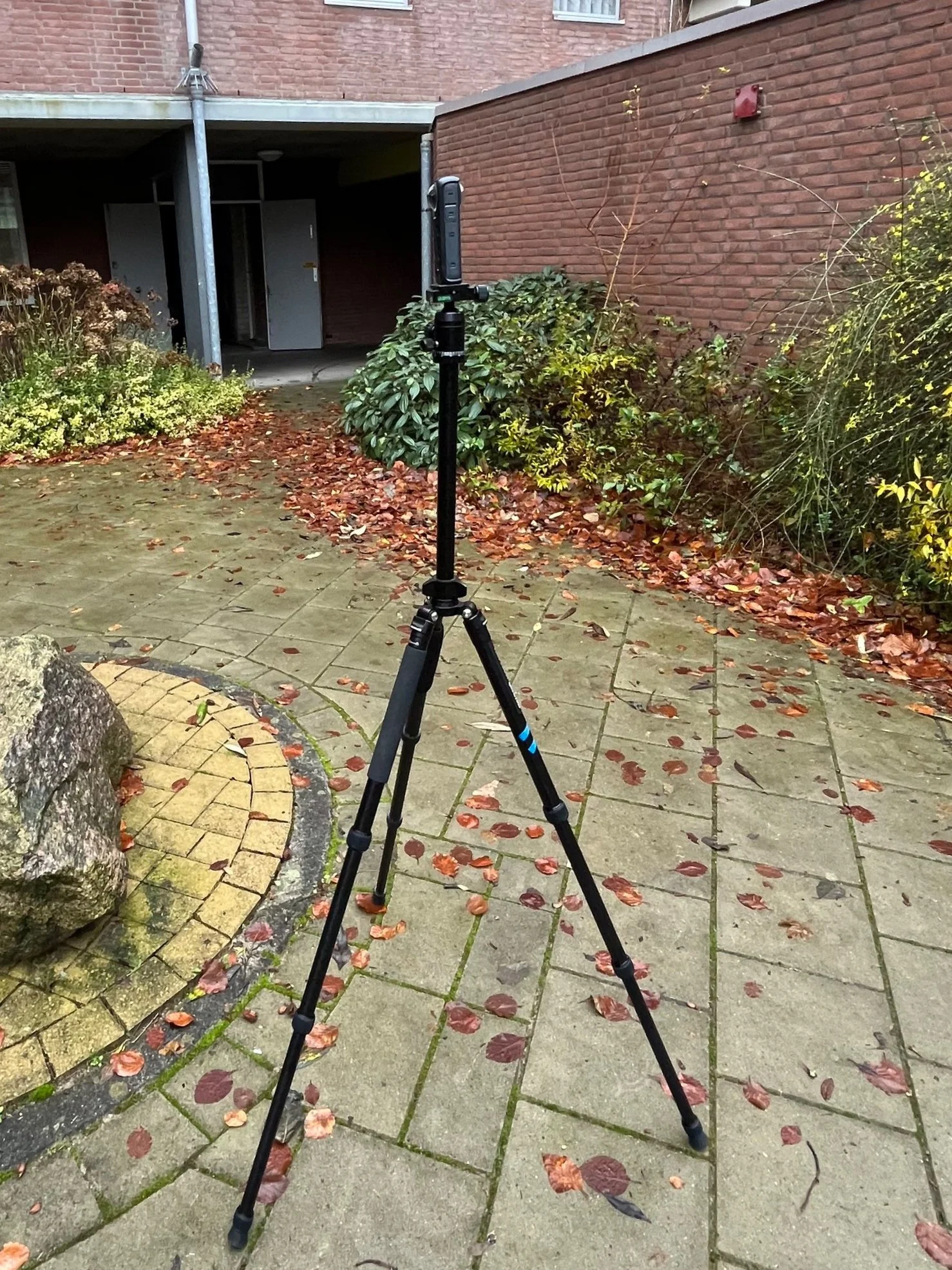 Een digitale camera op een statief staat op een betegeld pad, met herfstbladeren en struiken op de achtergrond.
