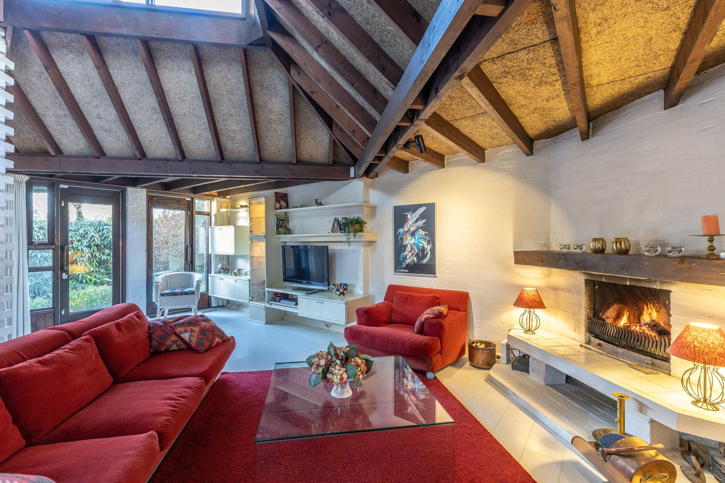 Woonkamer met rood bank en stoel, open haard met vuur, glazen tafeltje, wit muur, televisie en wandplanken met decoraties, grote ramen en deur naar tuin, houten plafond met dakbalken en plafondverlichting.