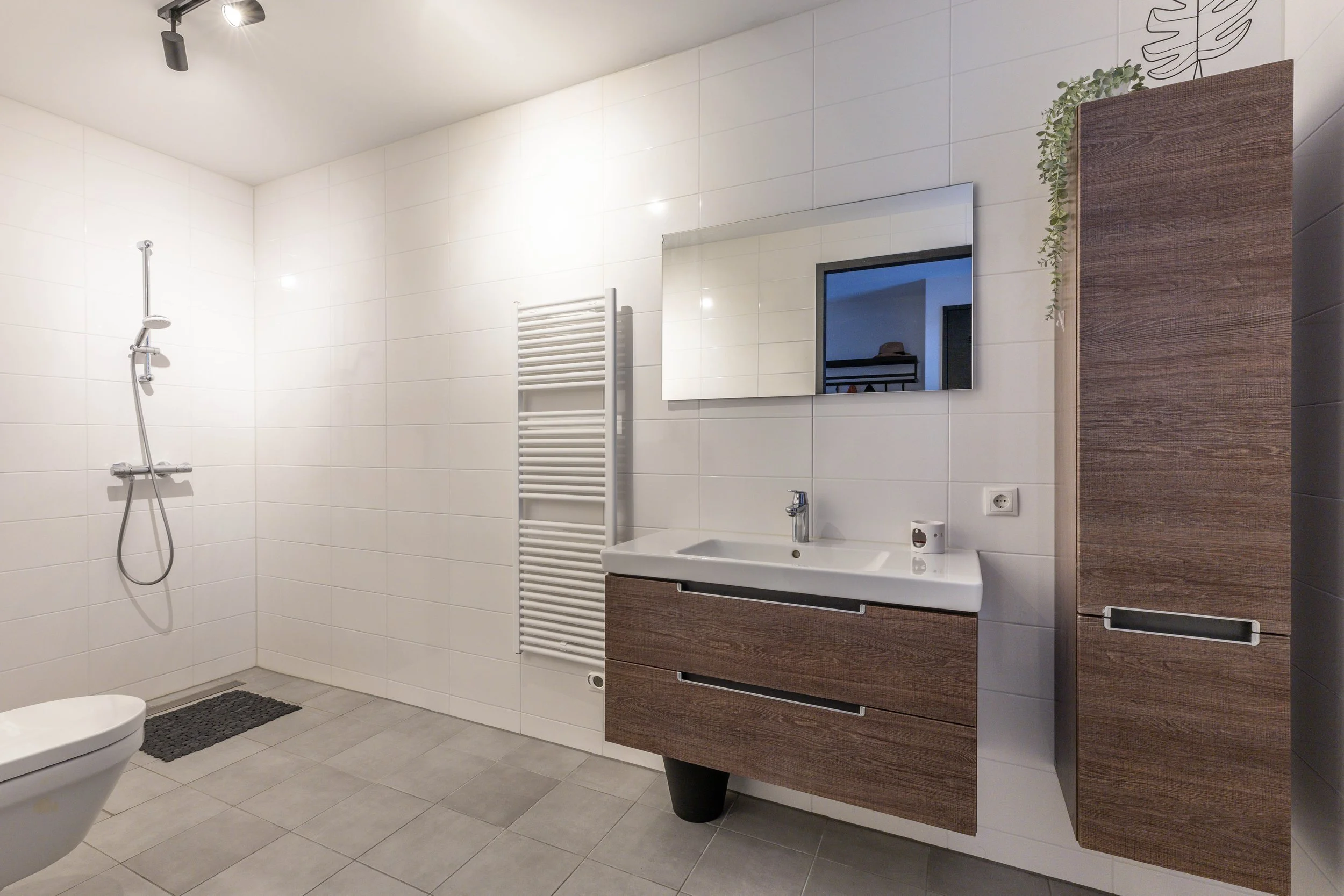 Een moderne badkamer met witte tegels, een vloer met grijze tegels, een spiegel, een houten badkamermeubel met witte wasbak, een handdoekradiator, een douche met een handdouche, een toilet, een houten kast en wat decoratieve planten.