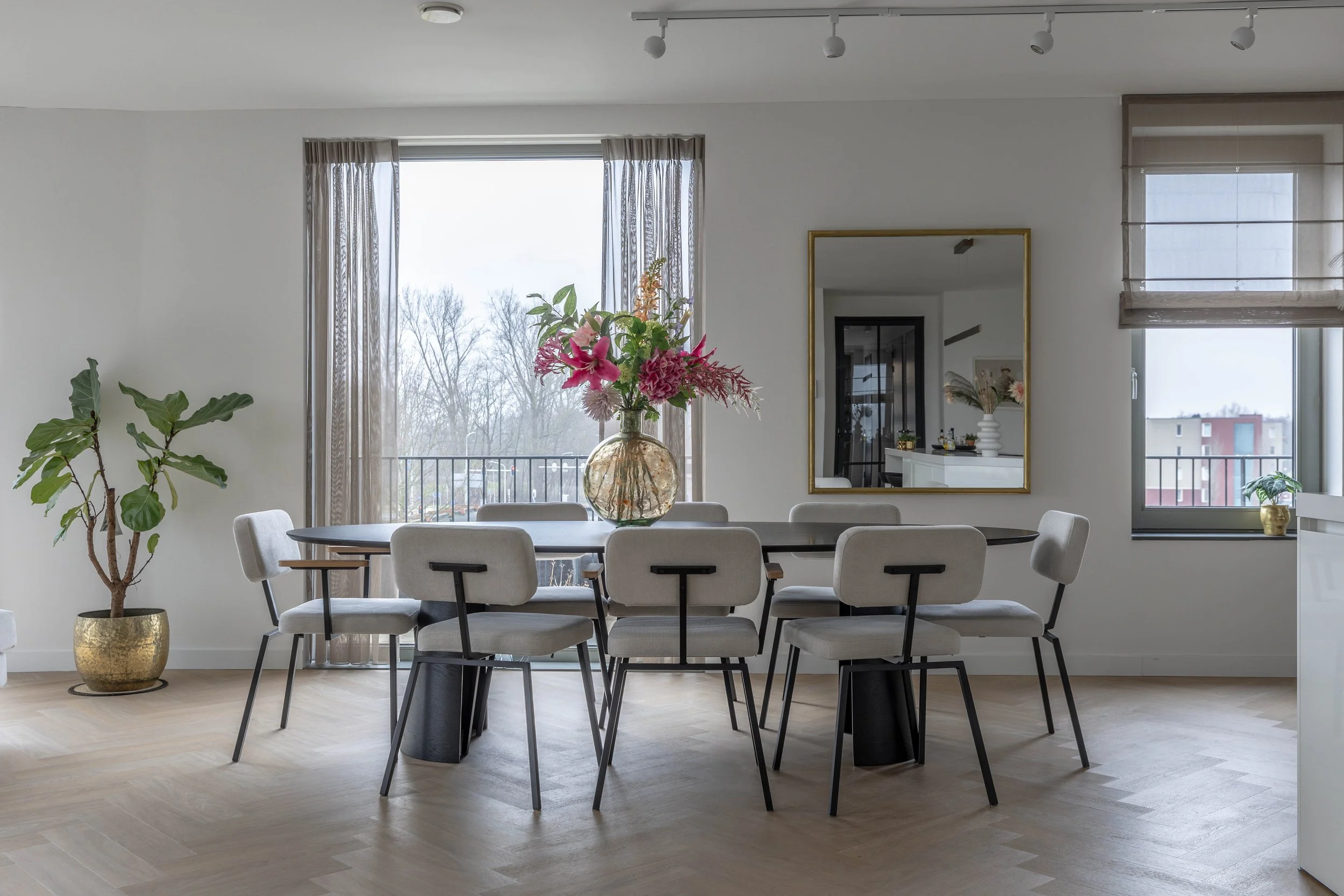 Eetkamer met een grote tafel, acht witte stoelen, een transparante vaas met bloemen, een grote plant in een gouden pot, een spiegel aan de muur, grote ramen met uitzicht op een balkon en een woonruimte op de achtergrond.