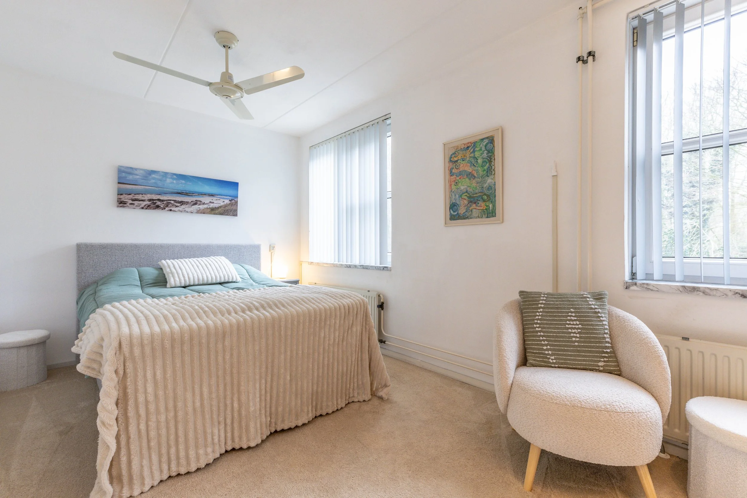 Een slaapkamer met witgekalkte muren, een bed met een lichtgrijze hoofdbord en een beige deken, een klein ronde stoel, een comfortabele stoel met een groene kussen, twee ramen met verticale jaloezieën, enkele kunstwerken aan de muur, en een plafondve