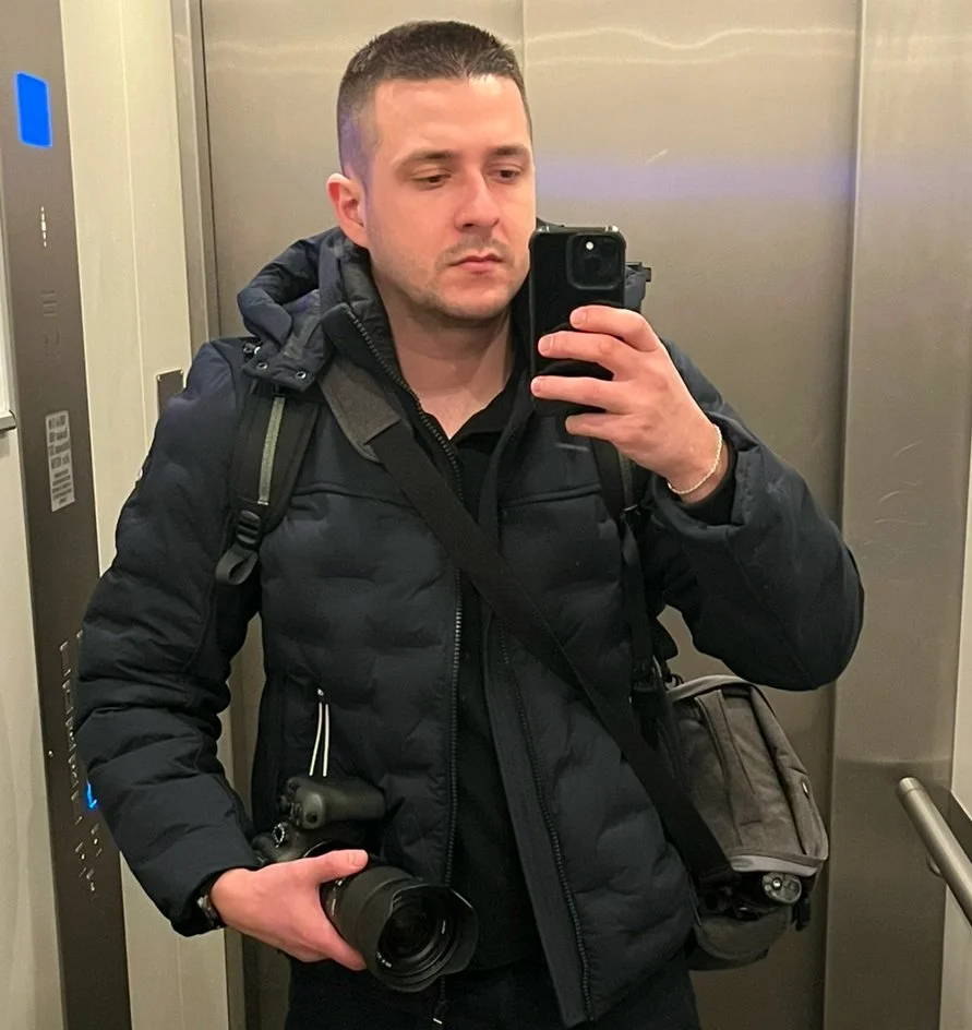 Man in een zwarte jas neemt selfie in een lift, hij houdt een camera in de hand en draagt een rugzak.