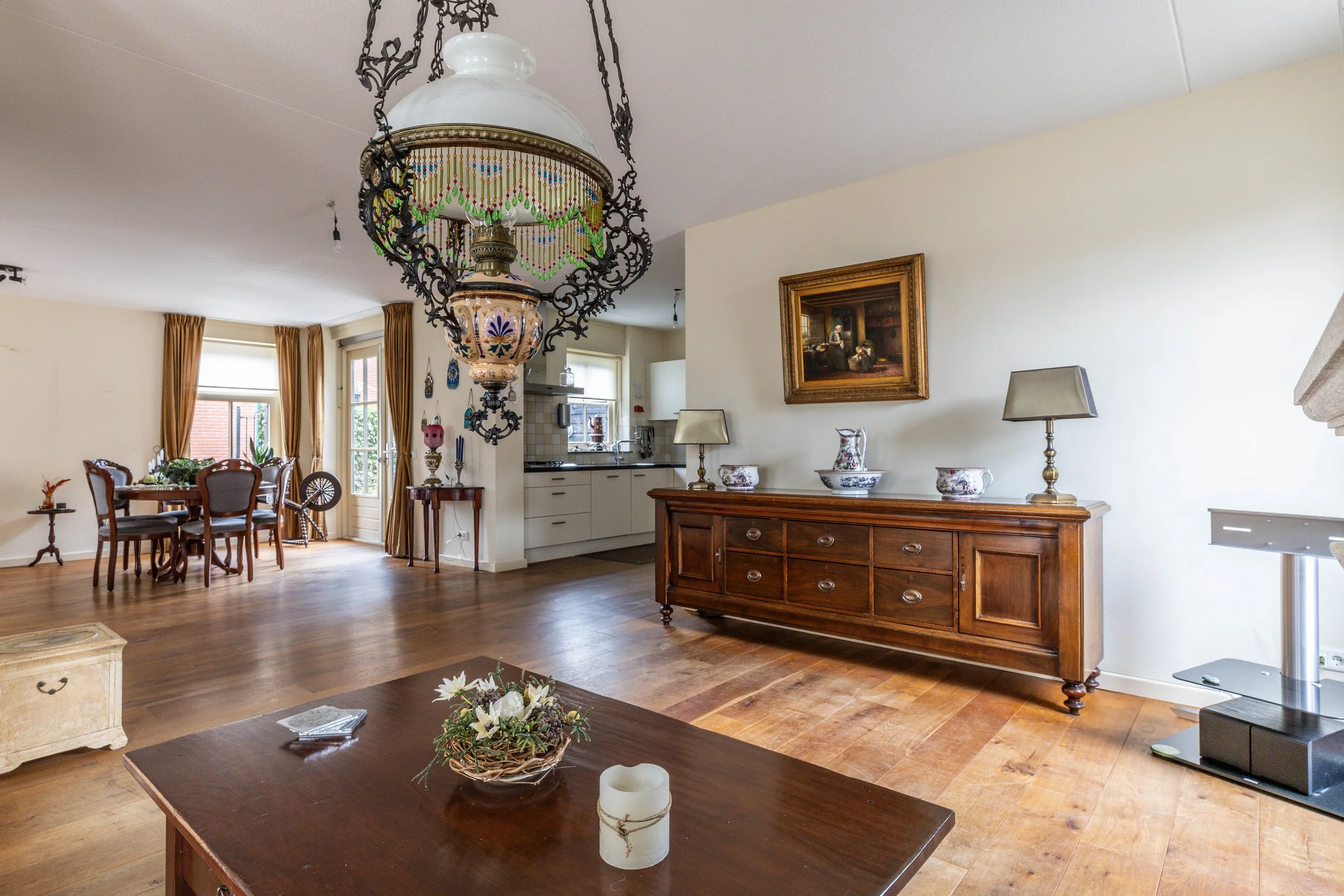 Woonkamer met houten vloer, antieke houten kast met decoratieve vazen en lampen, ronde eettafel met stoelen, grote lichtkoepellamp met beaded hangdecor, kunstwerk aan de muur, toegang tot keuken en balkon met deuren en gordijnen.