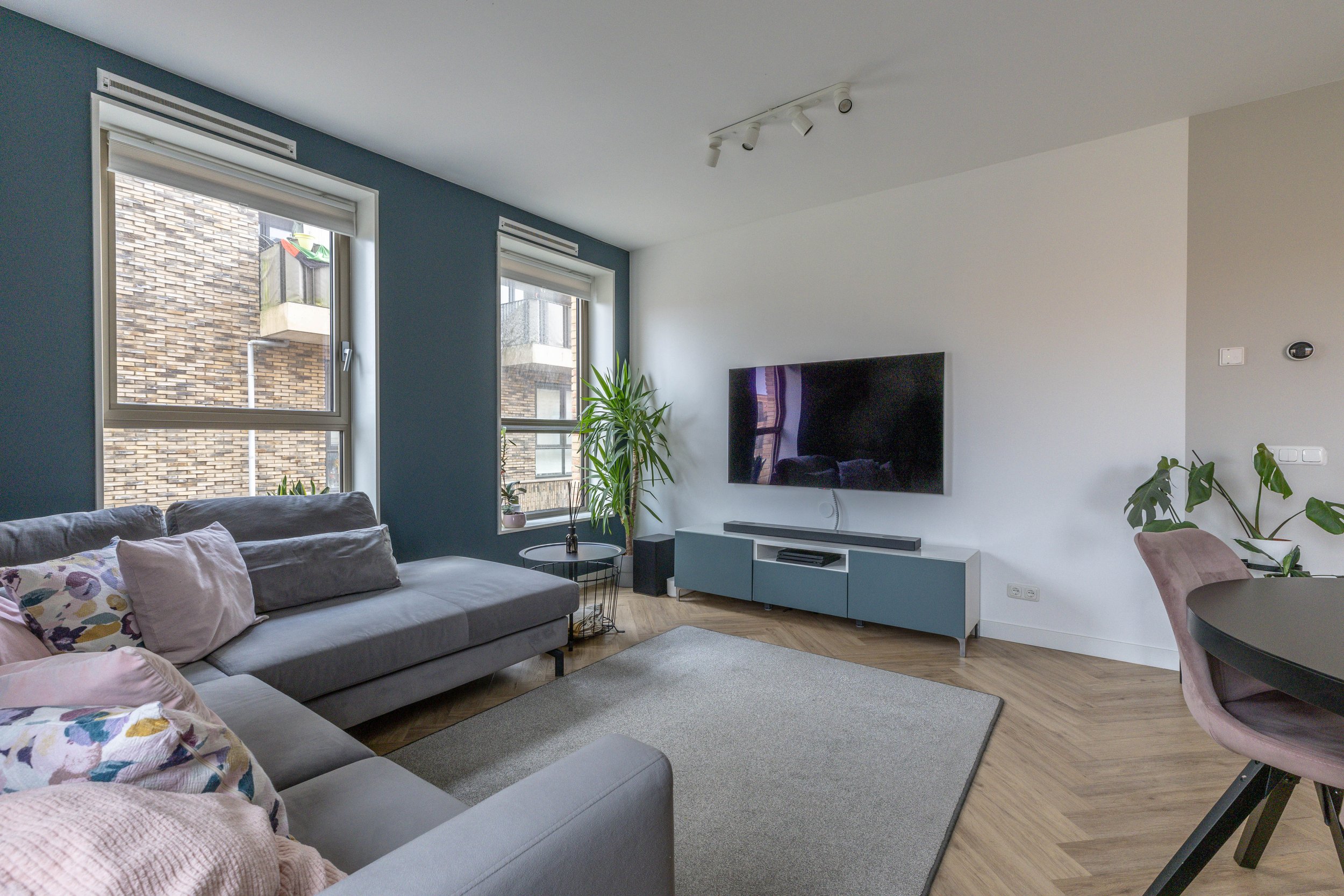 Modern woonkamer met grijze bank, tv aan de muur, planten en grote ramen met uitzicht op een bakstenen gebouw.