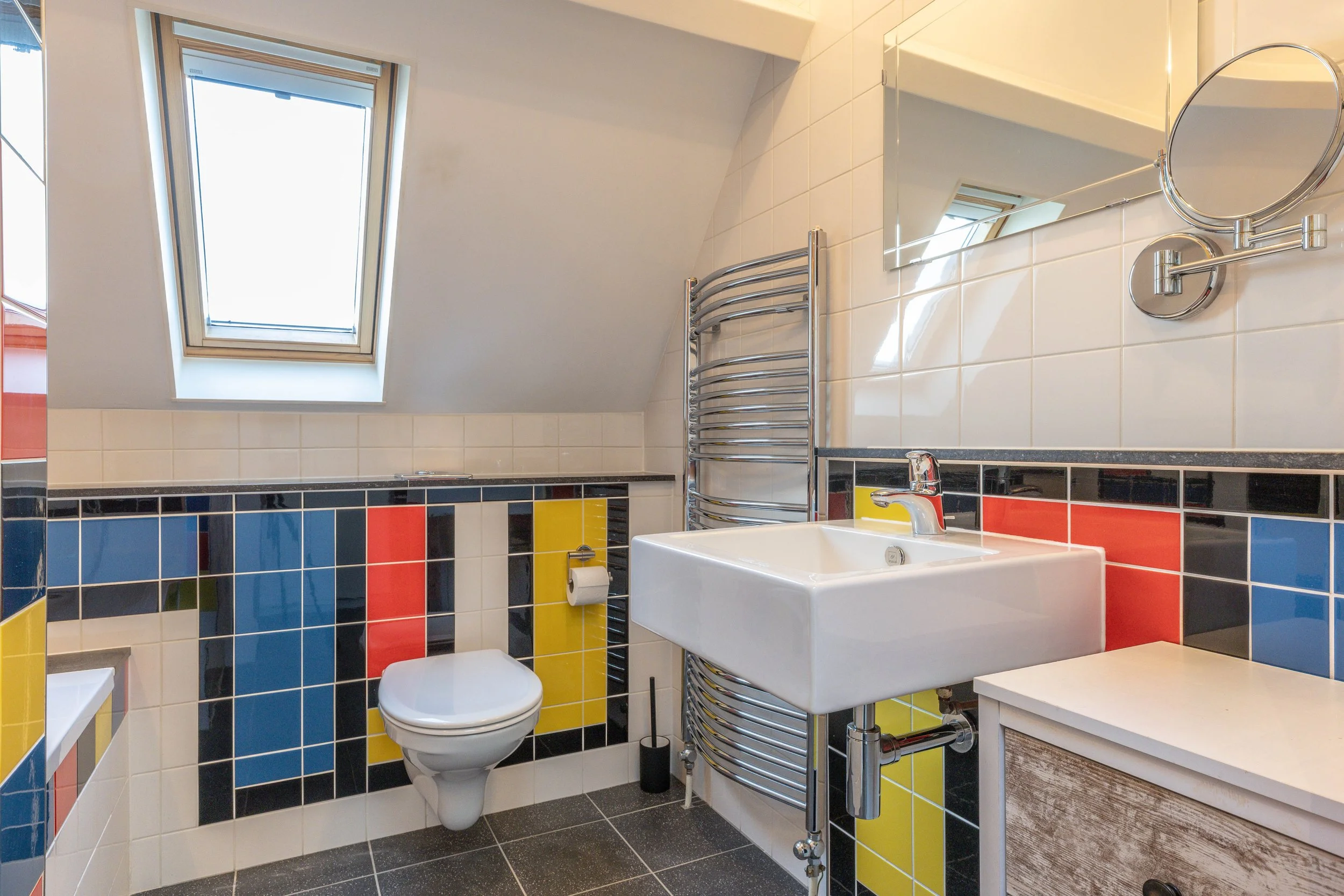 Kleine badkamer met een schuin dak en een dakraam, betegeld met kleurrijke tegels in blauw, rood, geel en zwart, inclusief een muurwasbak, spiegel, handdoekradiator en toilet.