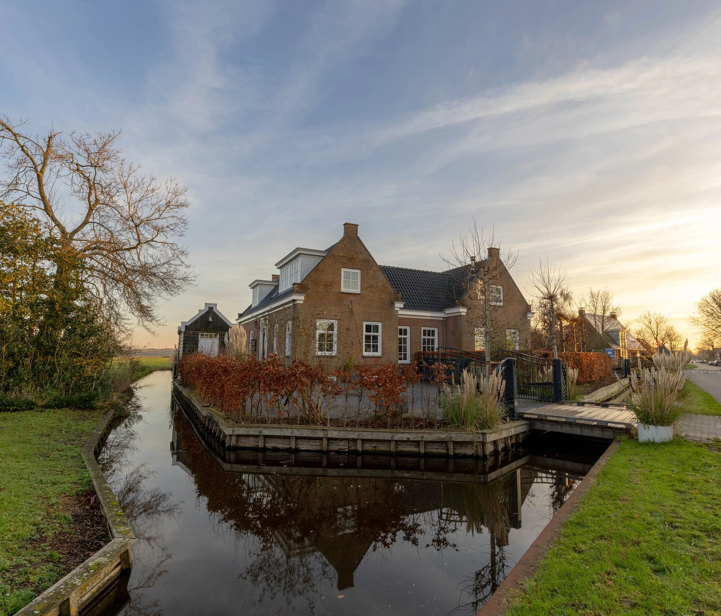 Een huis omringd door water met een kleine houten brug erbij, in een landelijke omgeving bij zonsondergang.