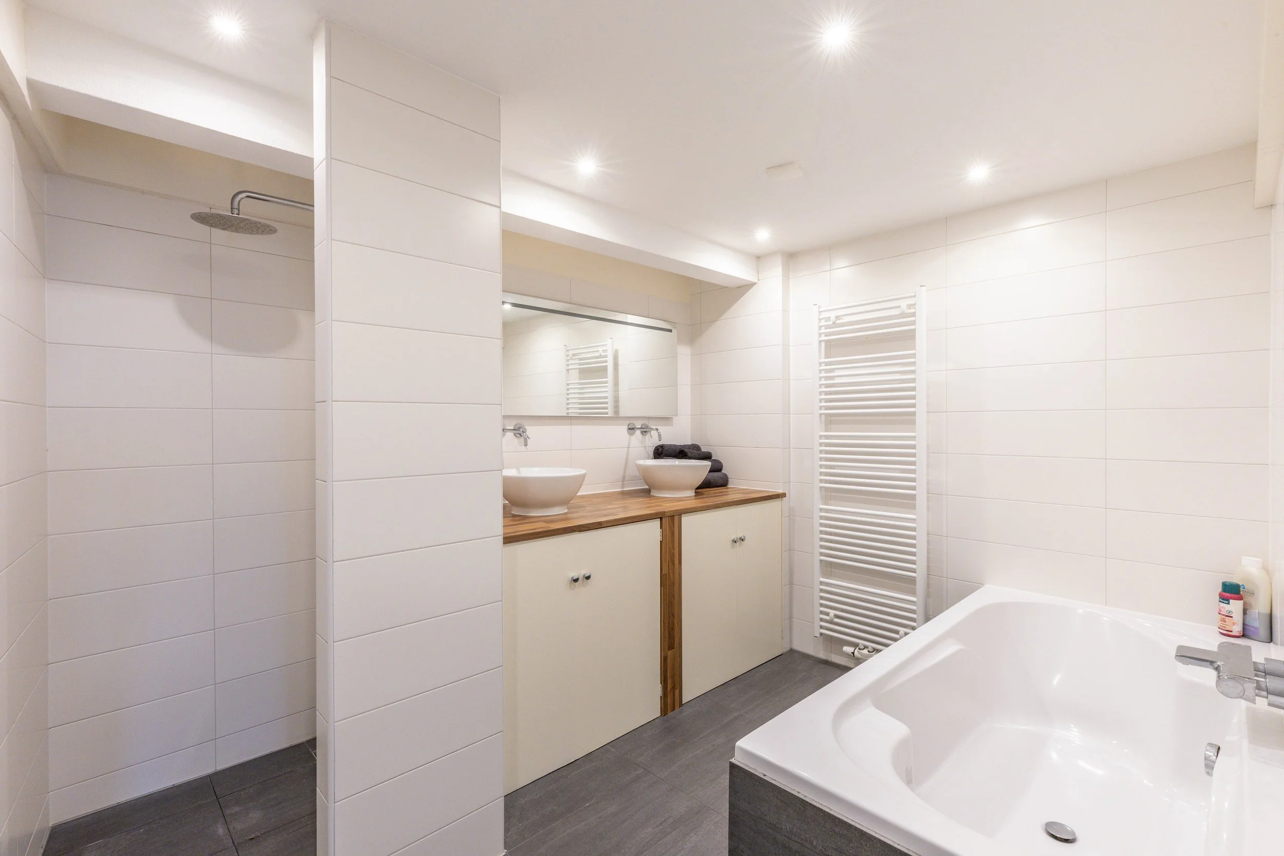 Moderne wit betegelde badkamer met inloopdouche, dubbele wastafel met ronde waskommen, grote spiegel en handdoekradiator.