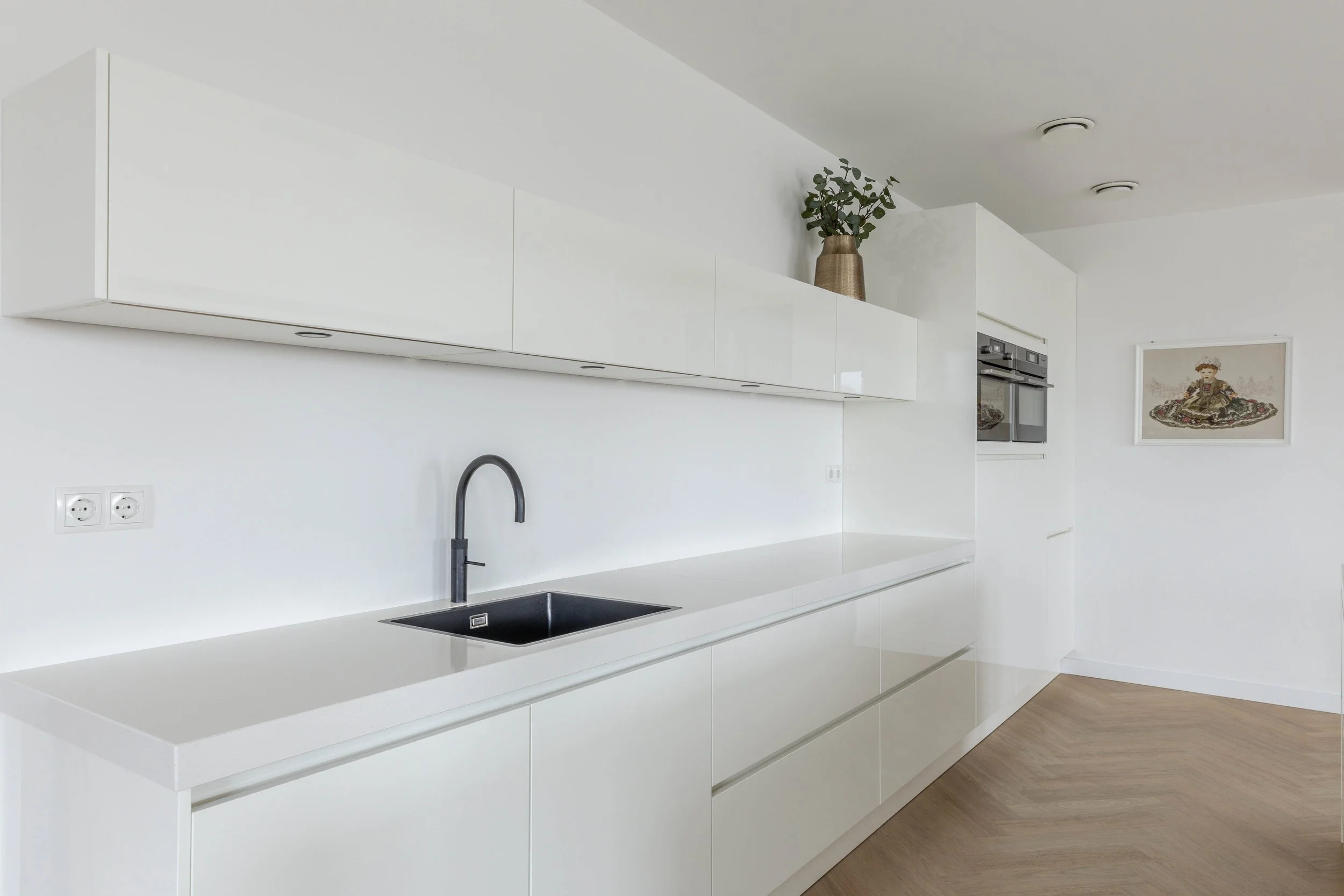 Minimalistische witte keuken met zwarte gootsteen en kraan, bovenkasten, een plant in een vaas op de bovenkast, en een kunstwerk aan de muur.