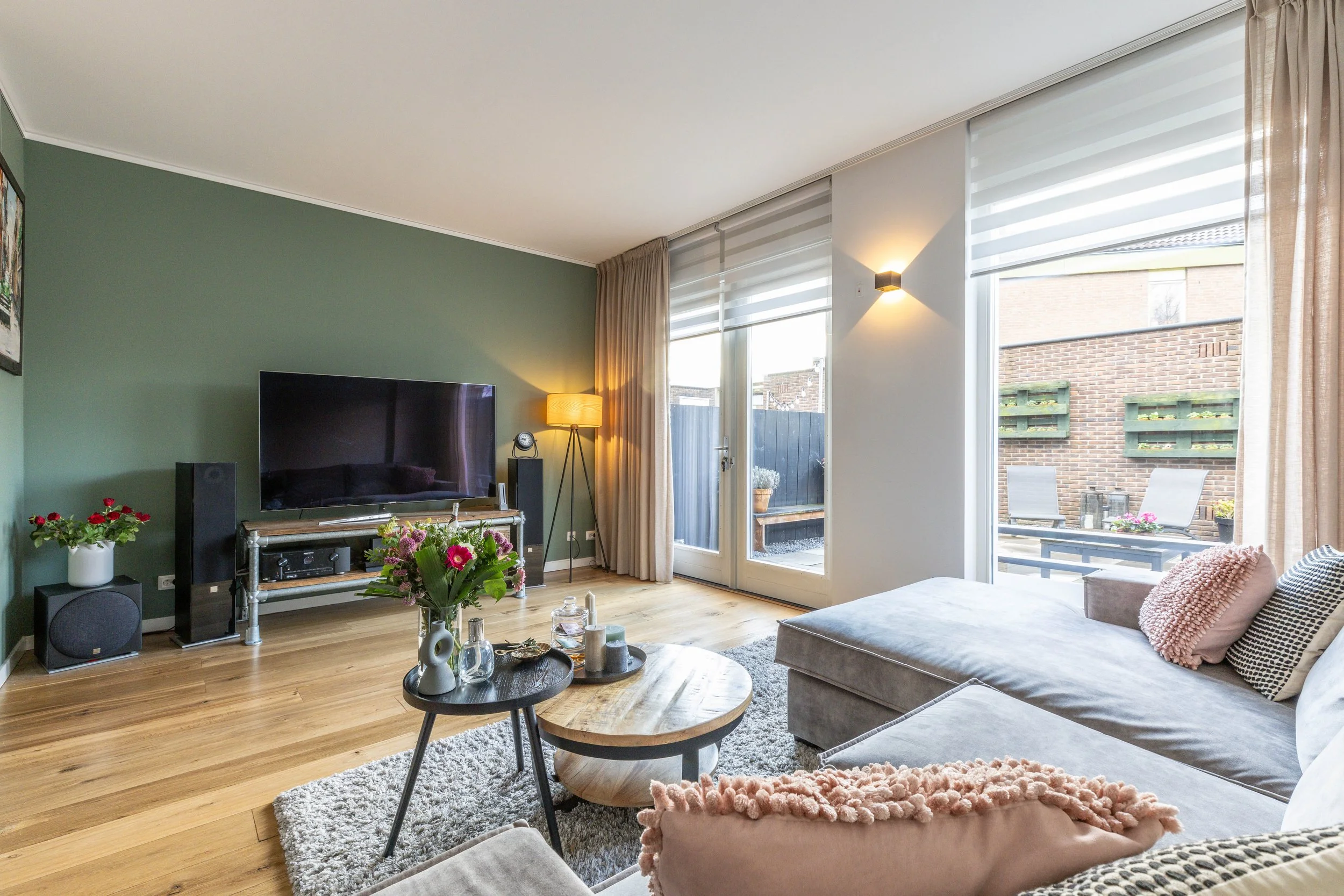 Lichte woonkamer met grote ramen en schuifdeur naar tuin, groene muur, beige bank met kussens, tv op metalen standaard, kleine bijzettafel met decoraties, houten vloer, terras met tuinmeubilair buiten.