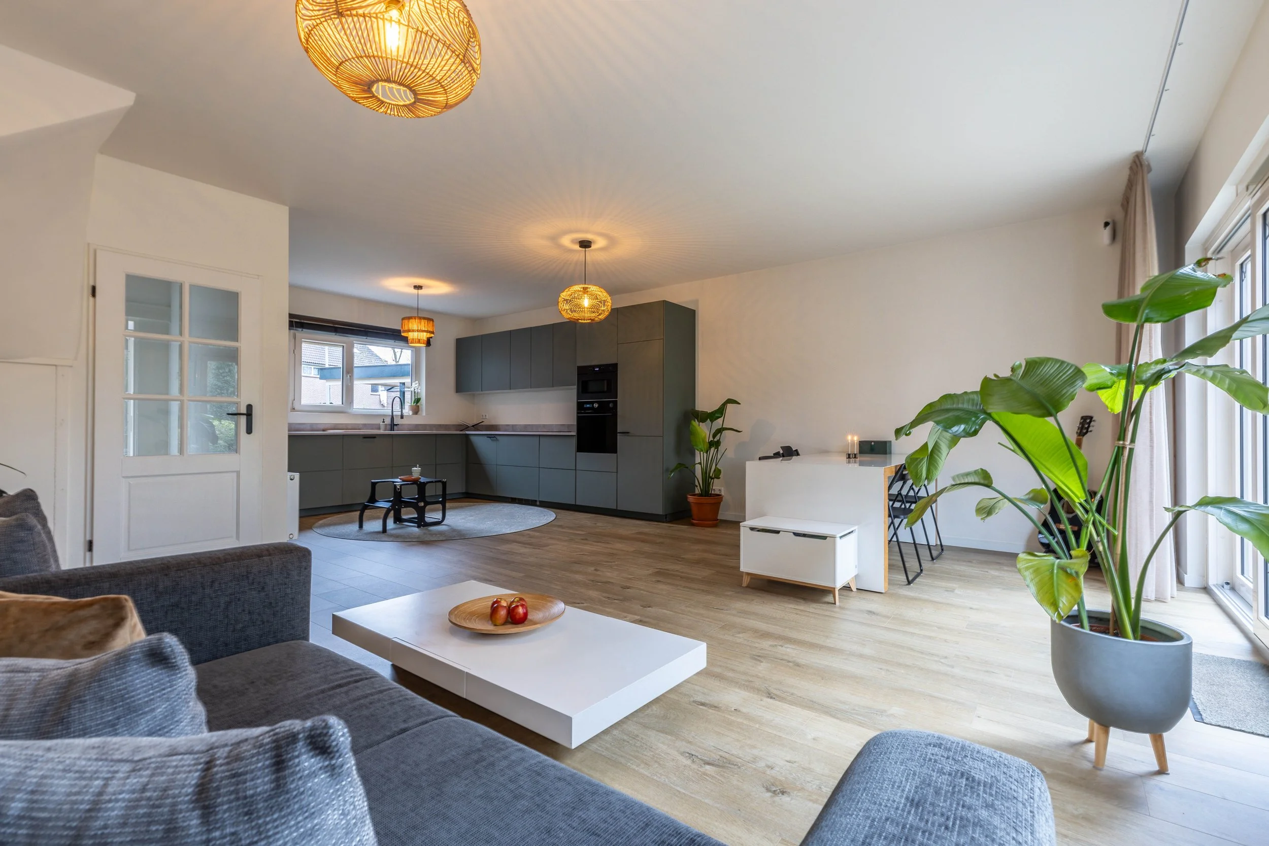 Een open woonkamer en keuken met natuurlijke lichtinval, grote groene planten, een wit/grijs plateau met appels en een grijze bank.