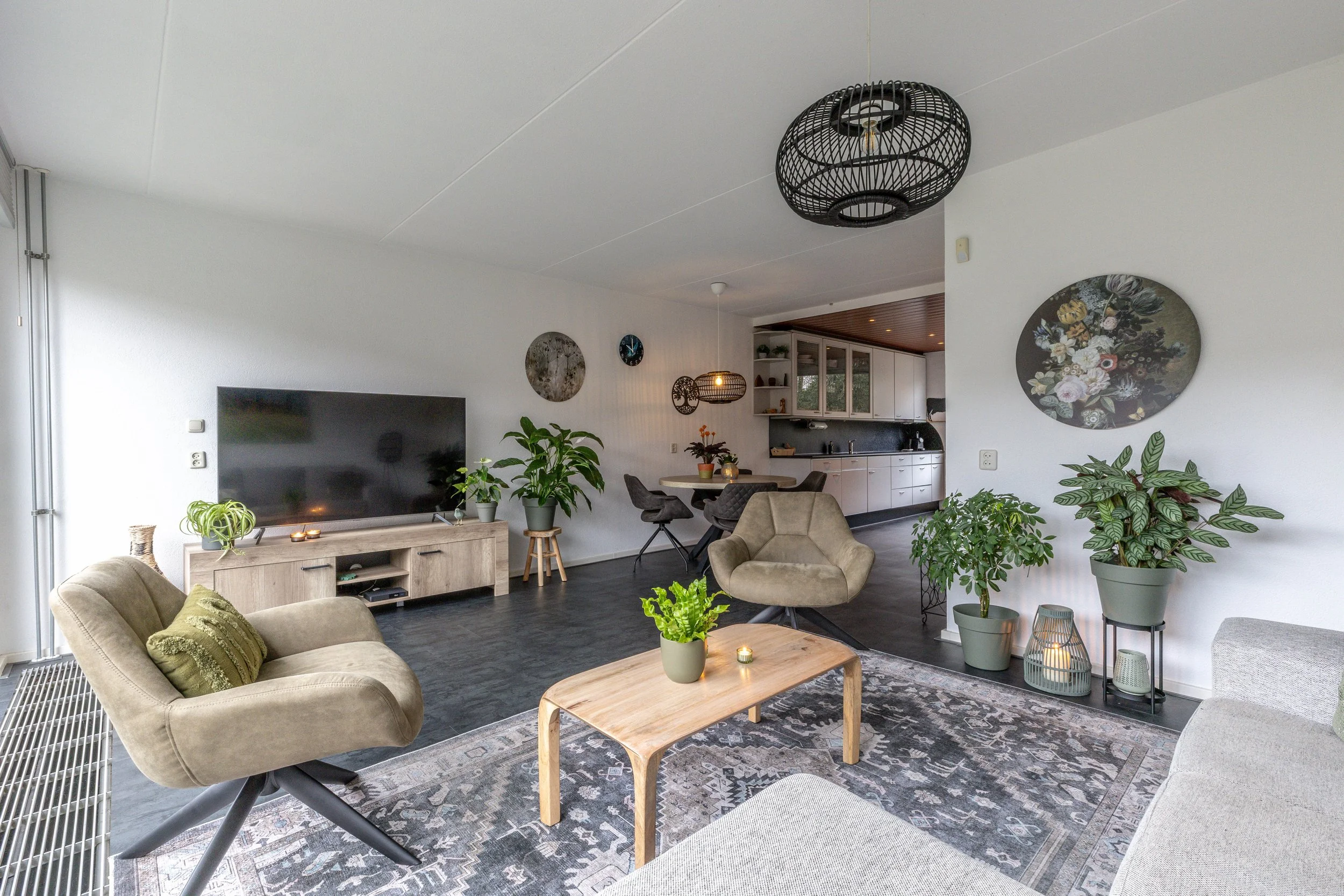 Een woonkamer met beige banken, een houten salontafel met planten en kaarsen, een grote flatscreen-tv op een witte tv-unit, diverse groene planten, wanddecoraties, een eethoek en een open keuken in de achtergrond. Moderne inrichting met natuurlijke t