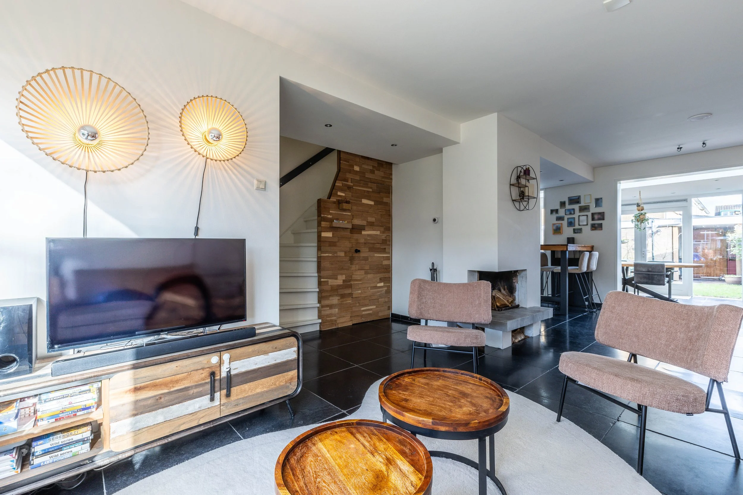 Modern woonkamer met een zwarte tv op een houten standaard, twee beige stoelen, twee kleine ronde houten tafels, een open haard, een muur met fotolijstjes, en een eetkamer met uitzicht op de tuin.