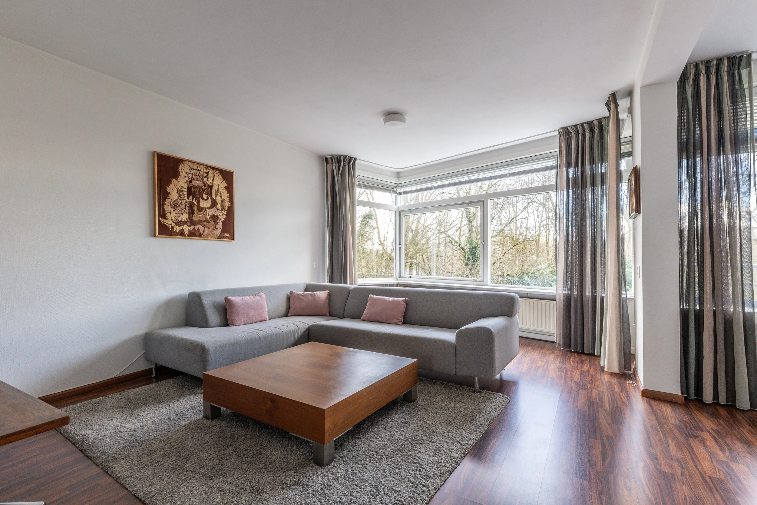 Woonkamer met grote hoekbank, houten salontafel, vloerbedekking en grote ramen met gordijnen, uitzicht op bomen buiten.