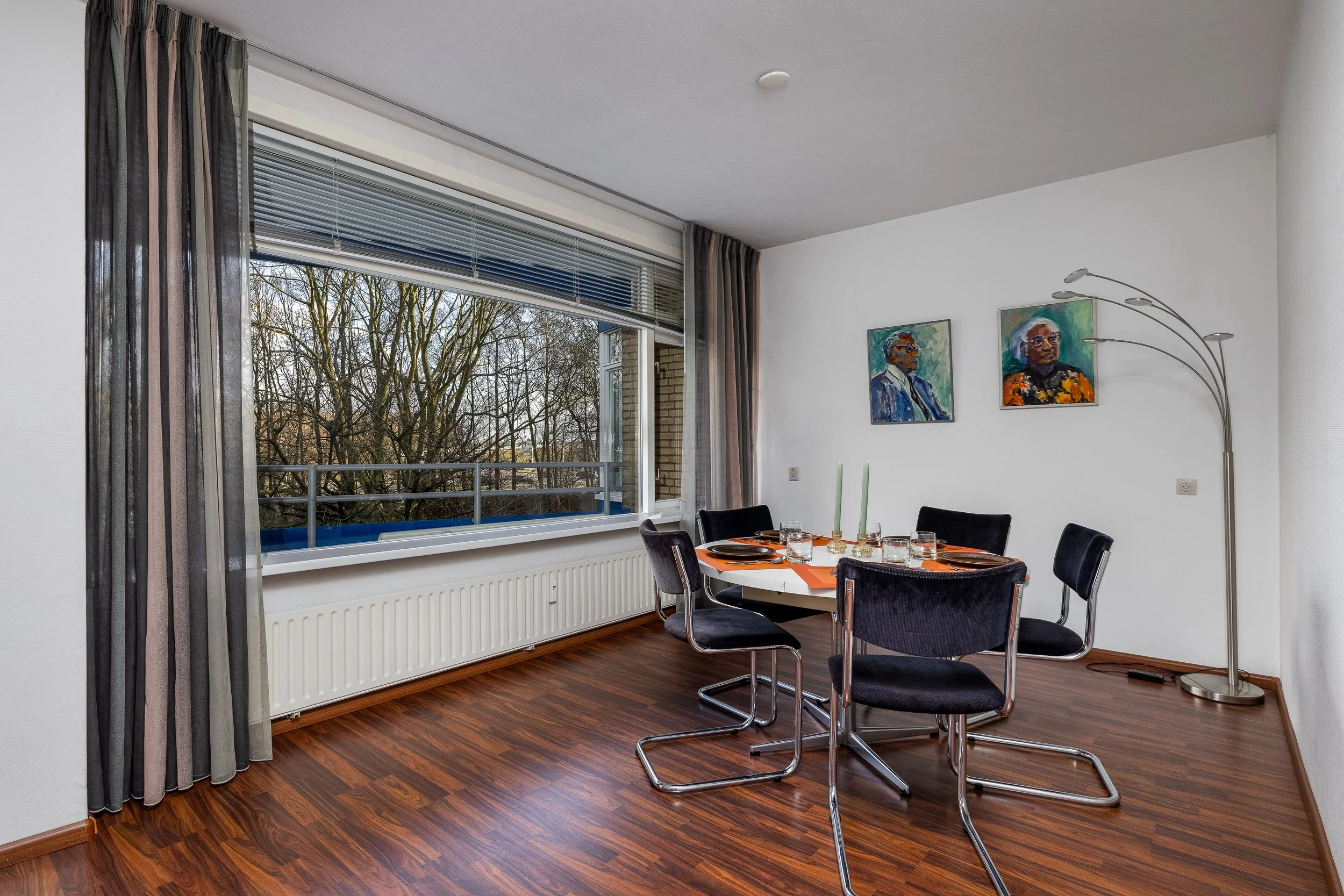 Eetkamer met ronde tafel, vier zwarte stoelen, grote open raam met uitzicht op bomen, twee schilderijen aan de witte muur, vloerlamp met meerdere armen, houten vloer, gedekte tafel met kaarsen en glazen.