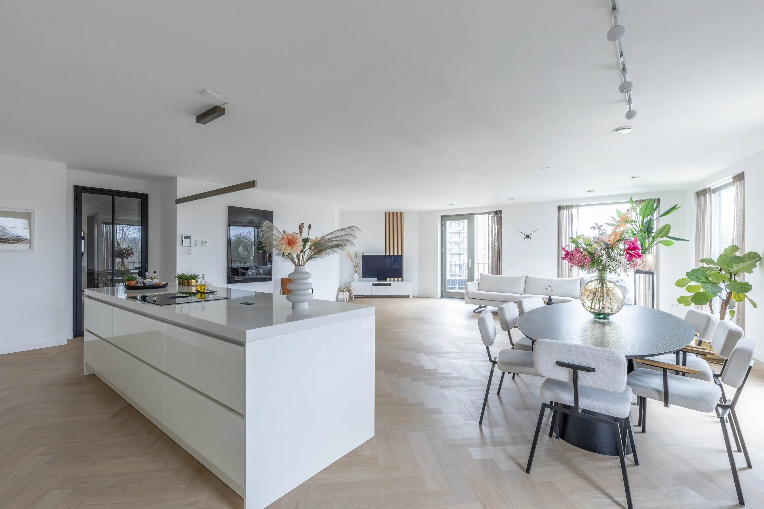 Open plan woonkamer en keuken met witte muren, licht houten vloer, ronde eettafel met acht stoelen, grote vensters met beige gordijnen, planten en bloemen in vazen, tv en balkon in de achtergrond.