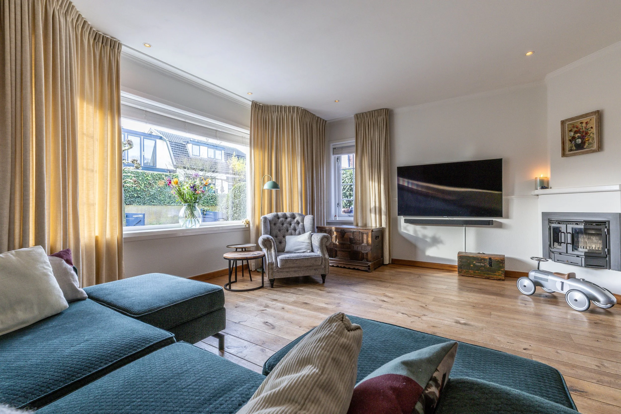 Woonkamer met grote ramen, beige gordijnen, een bank, een armstoel, een bloemenvaas, een tv aan de muur, houten vloer, en een kinderauto.