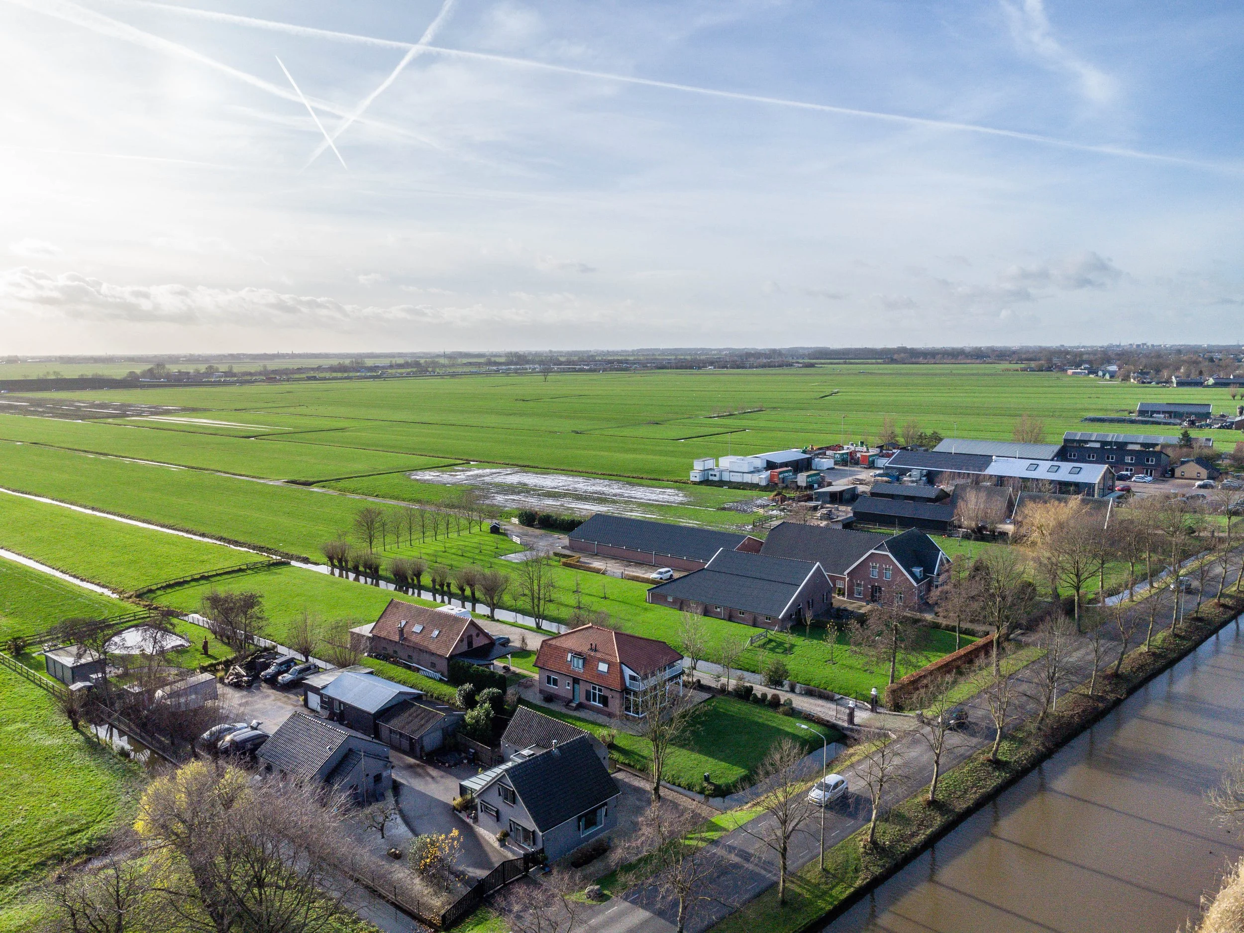 Luchtfoto van een polderlandschap met boerderijen, huizen en weilanden, gescheiden door sloten en wegen, met een rivier aan de rechterkant en een open hemel met enkele vliegerstrepen.