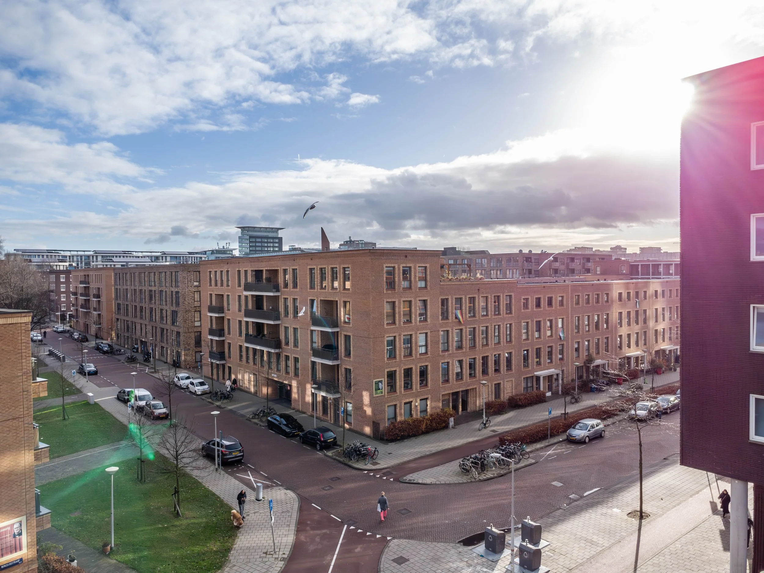 Uitzicht op een stedelijk gebied met bruine appartementencomplexen, een straat met geparkeerde auto's, fietsen, en voetgangers, onder een deels bewolkte blauwe hemel met zonlicht.