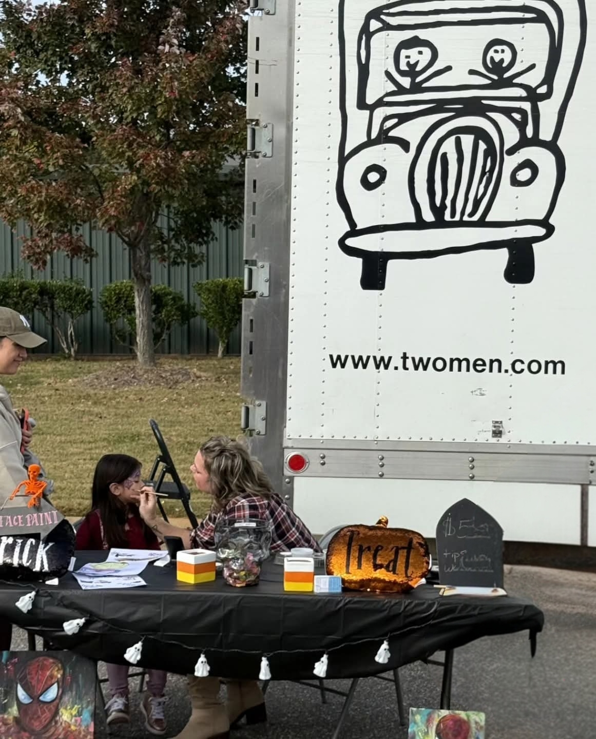 Woman painting a child's face at a Halloween-themed outdoor event, with a table displaying Halloween decorations and treats, and a large truck with a simple black line drawing of a car and the website www.twomen.com in the background.