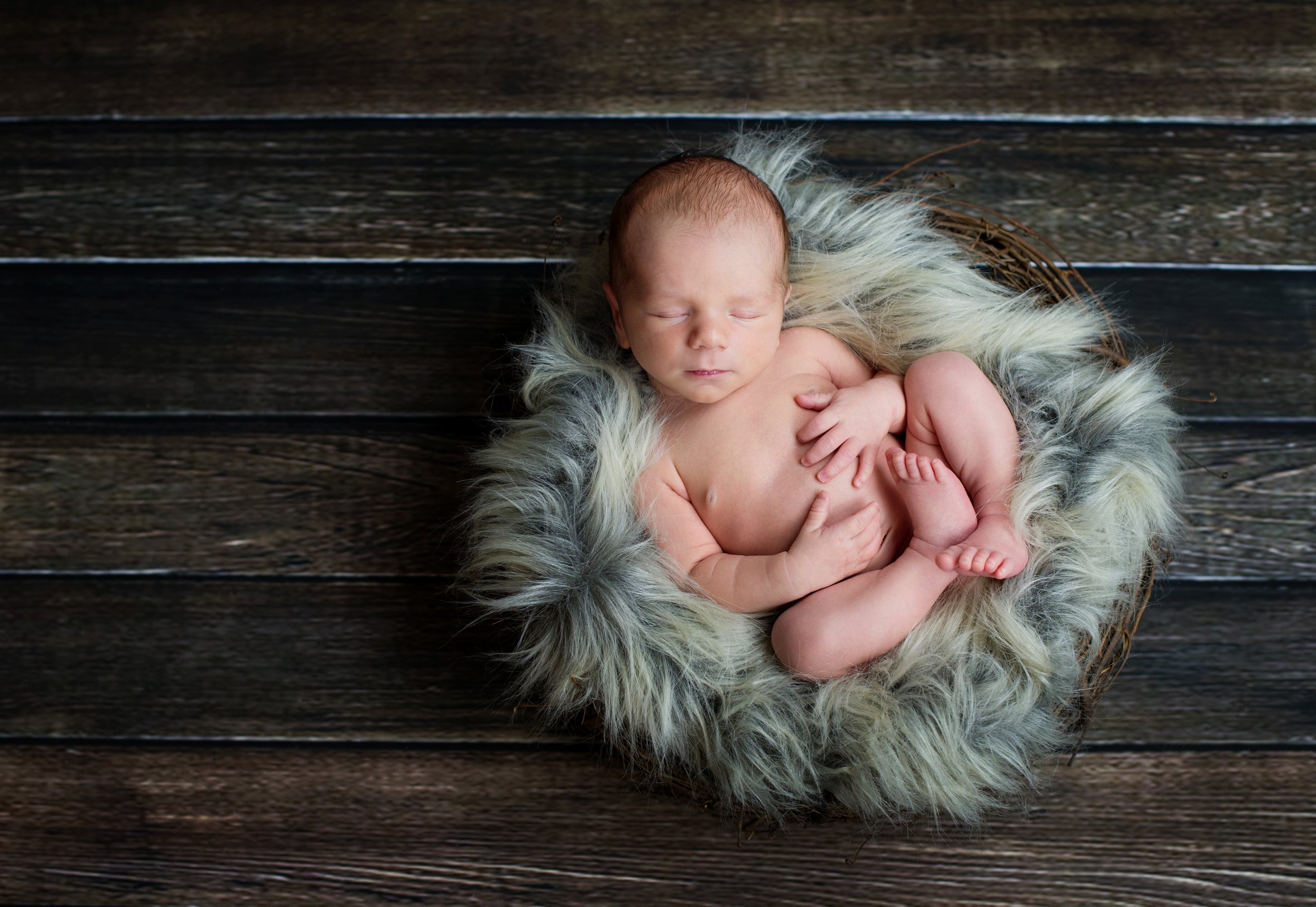 Newborn baby sleeping in a basket with soft fur lining on a wooden floor.