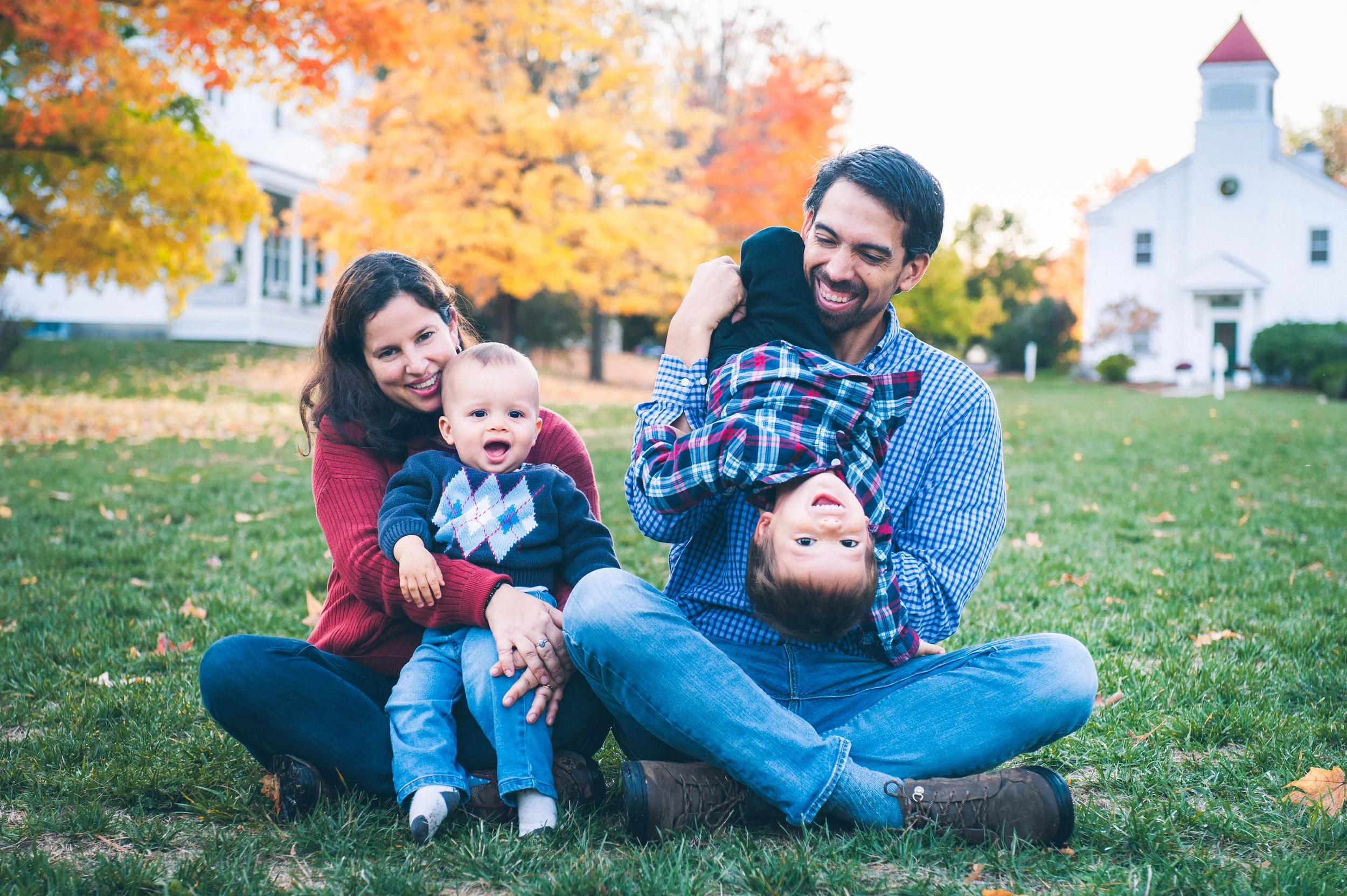 Family sitting on grass in autumn for a portrait, with colorful trees and a white house in background; two children and two adults smiling.