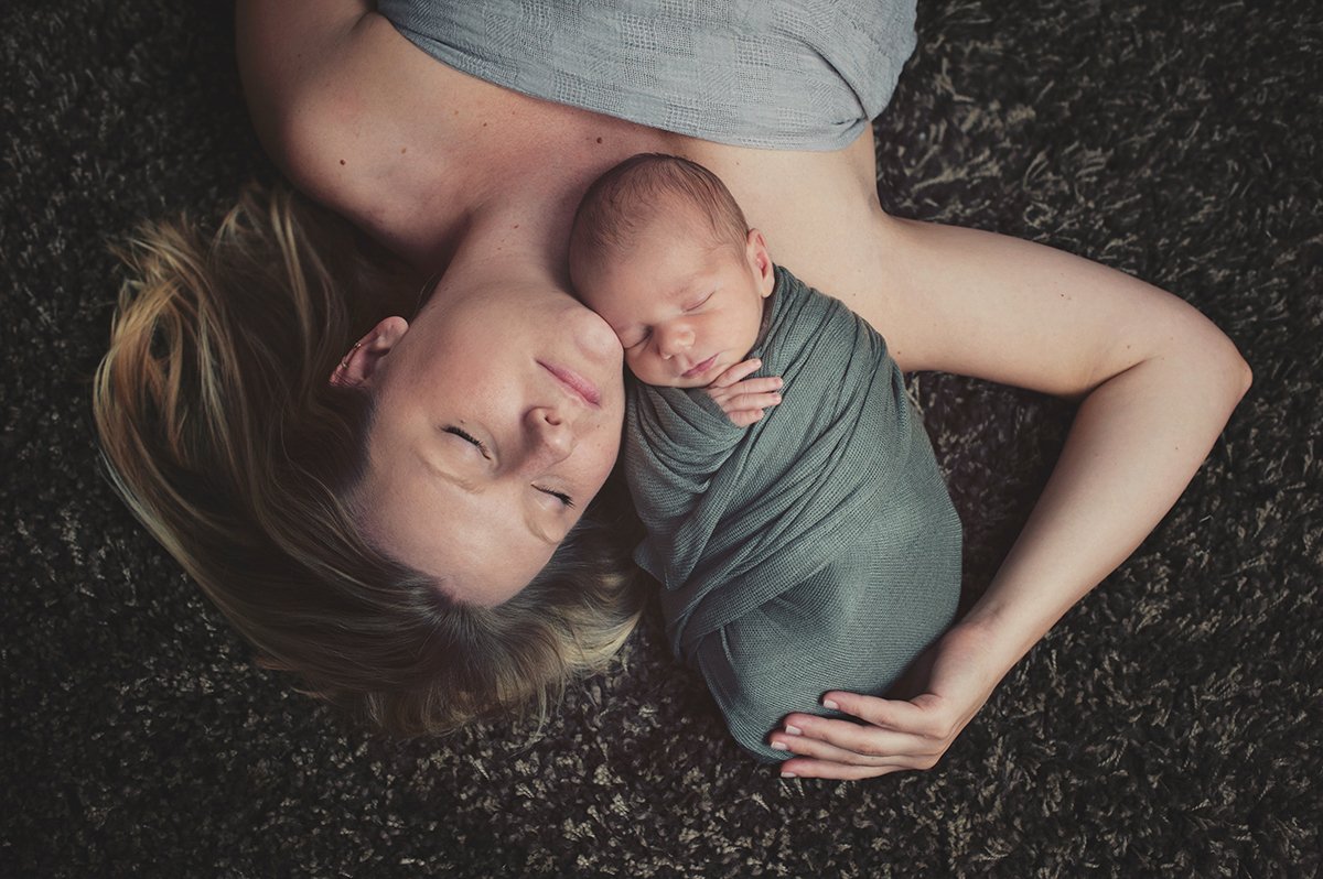 Mother and newborn baby lying on a dark carpet, with the baby wrapped in a blanket, both sleeping peacefully.