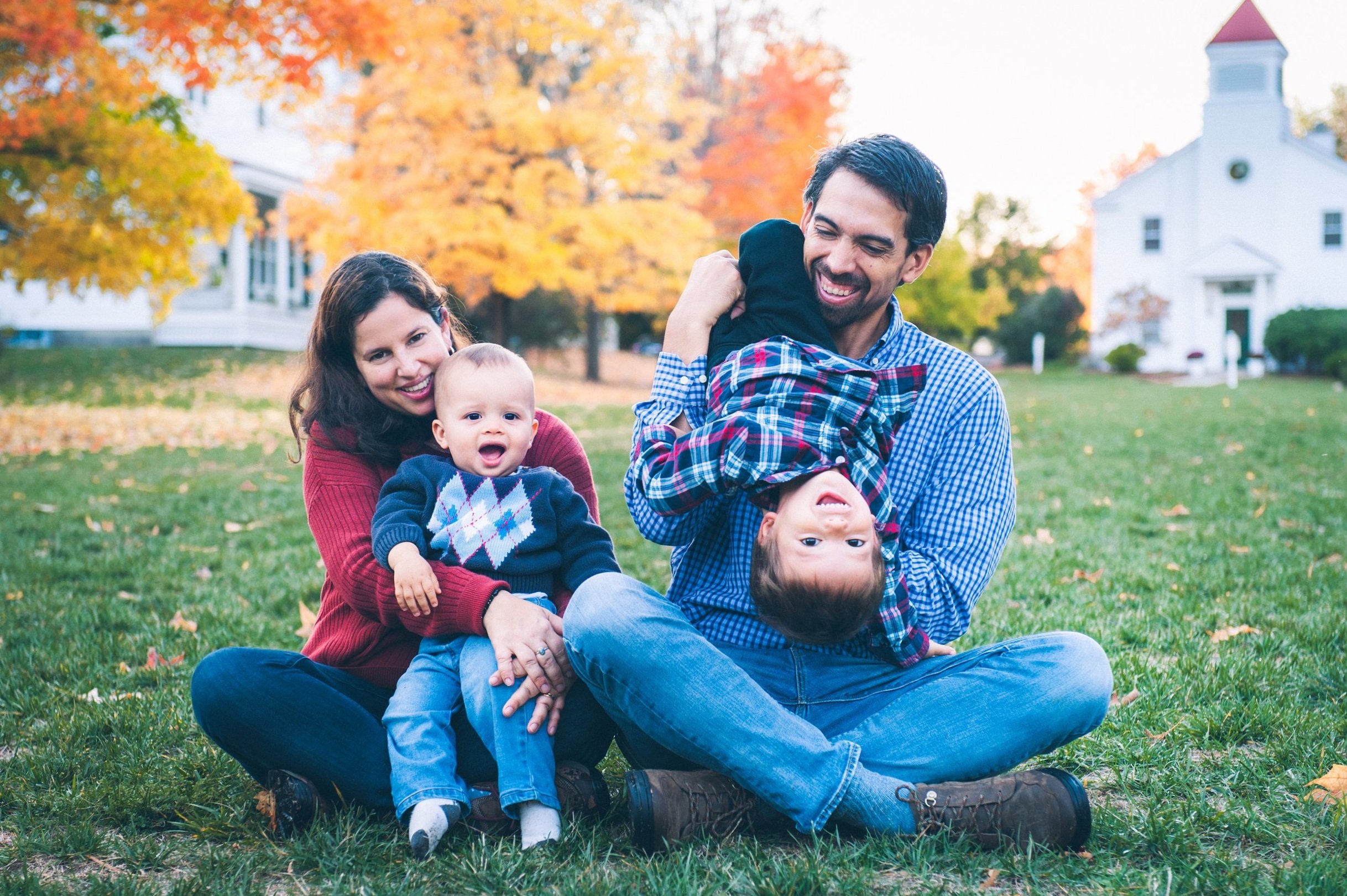 Portrait of a family sitting on grass with autumn leaves, mother holding baby, father playfully holding child upside down.